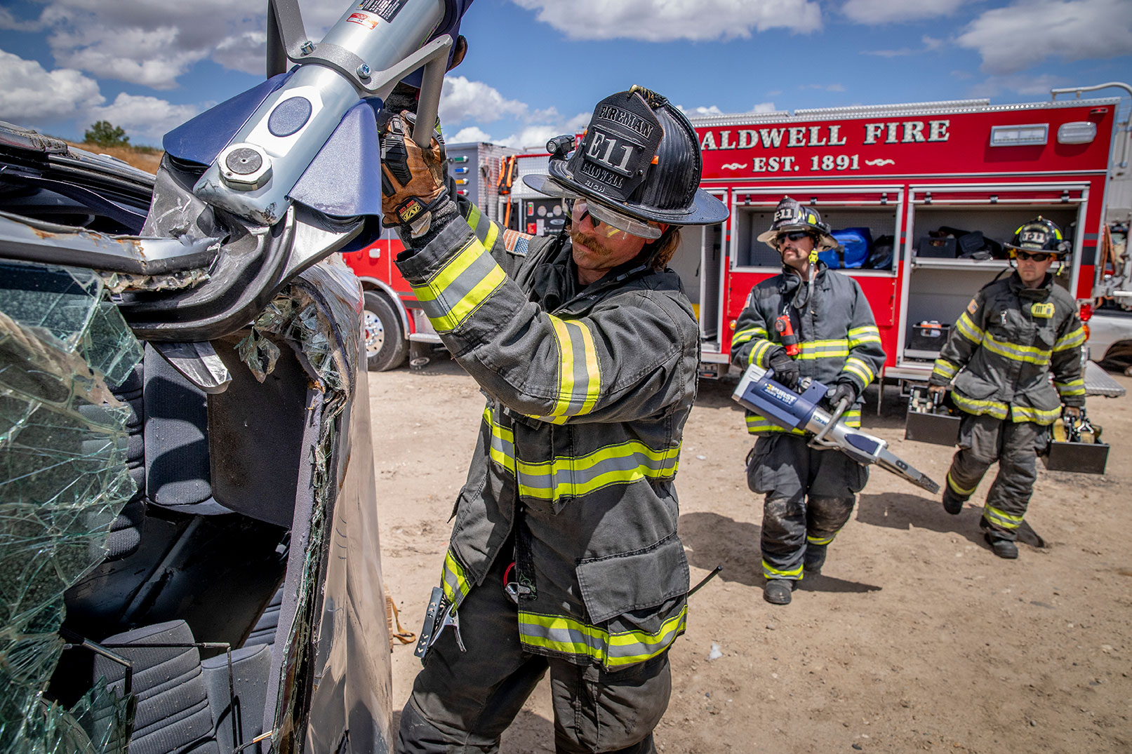 Three firefighters in turnout gear working with rescue equipment on a crashed car and a Heavy-Duty Rescue Pumper in the background.