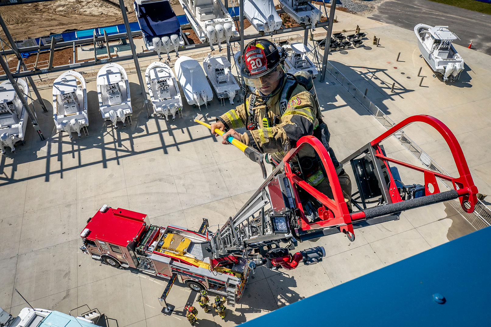 A firefighter in turnoutgear holding a long tool at the tip of an aerial ladder reaching toward a building and firefighters below.