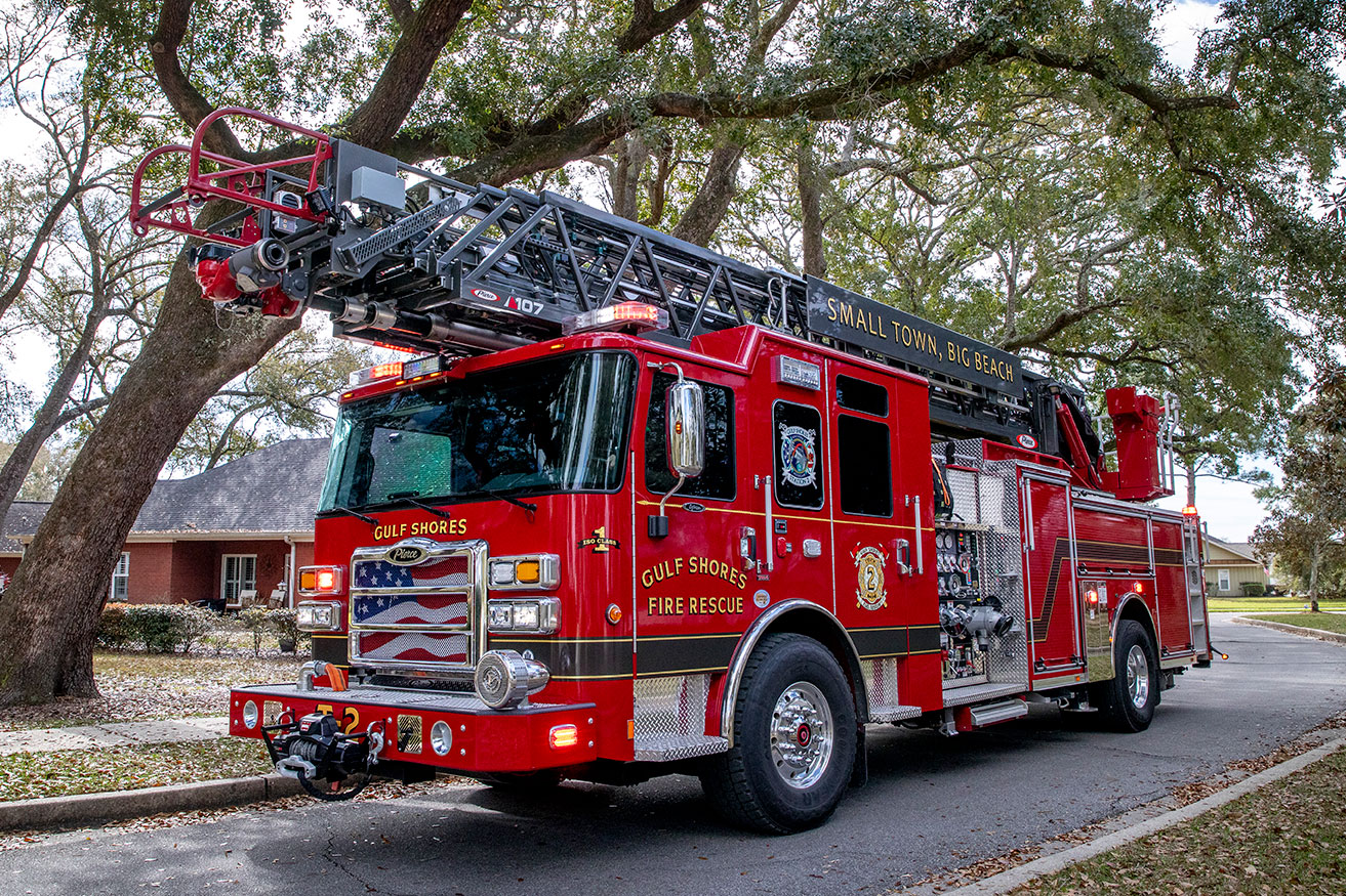 The front drivers side of an Enforcer Ascendant 107 aerial ladder driving down a road with overhanging green trees.