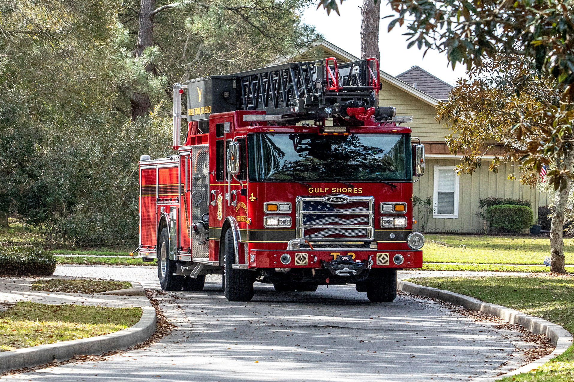 The front officers side of an Ascendant 107 aerial ladder driving on a windy road by green grass and trees.