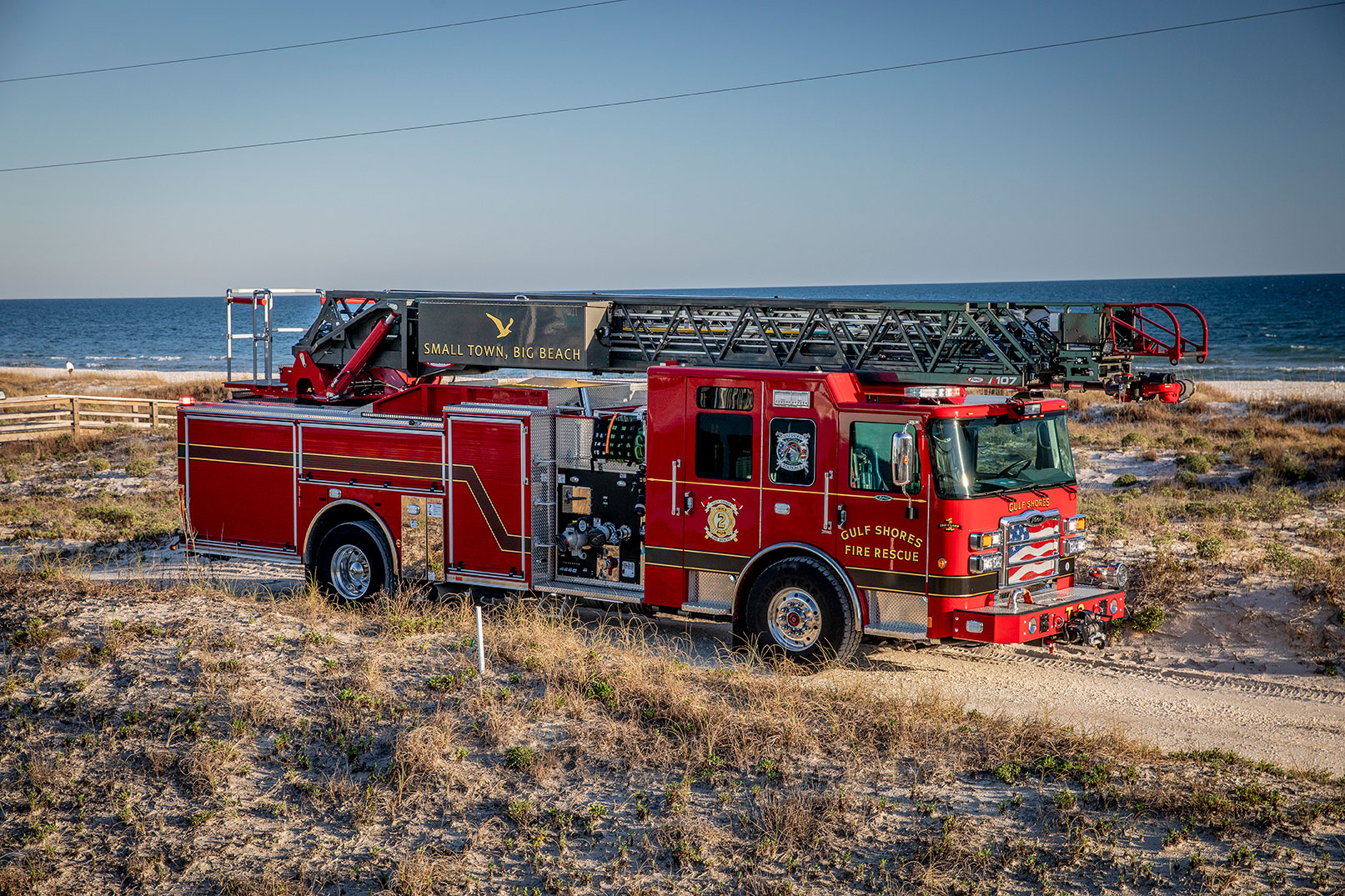 The officers side of an Enforcer Ascendant 107 Aerial Ladder driving on a beach next to the ocean.