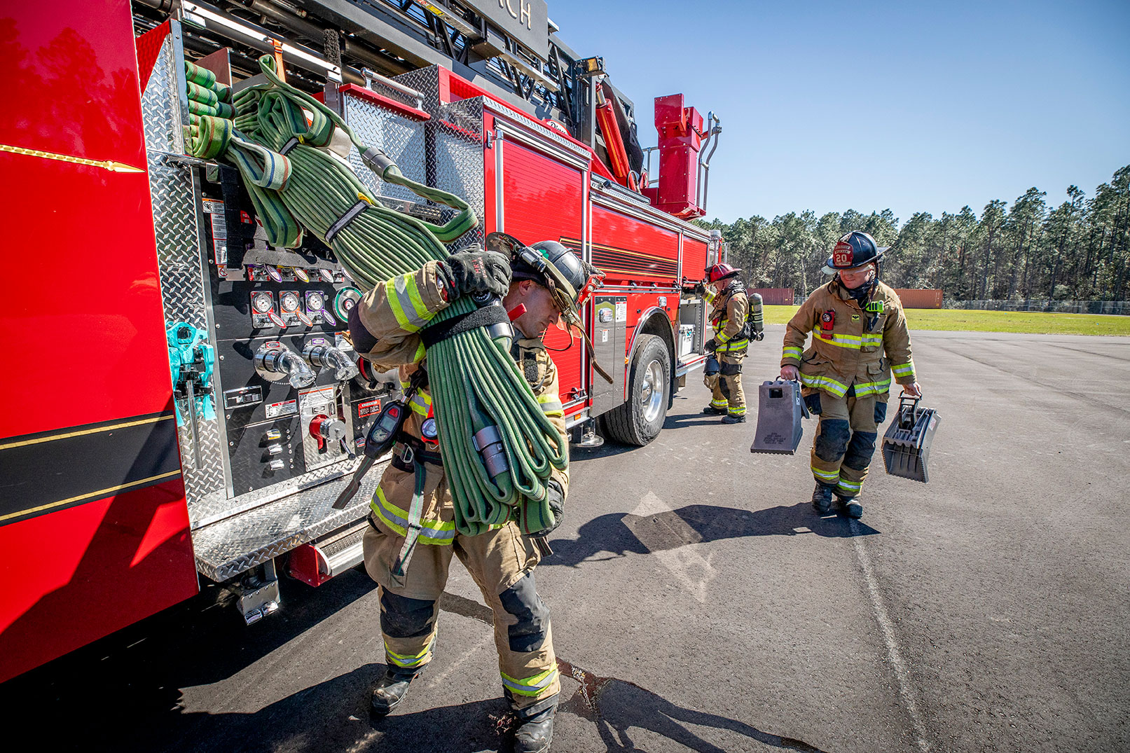 Three firefighters in turnout gear on the drivers side of a fire truck grabbing fire hose and wheel chocks.