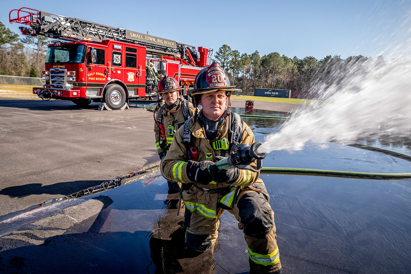 Two firefighters in turnout gear kneeling on the ground spraying water from a fire hose from a fire truck.
