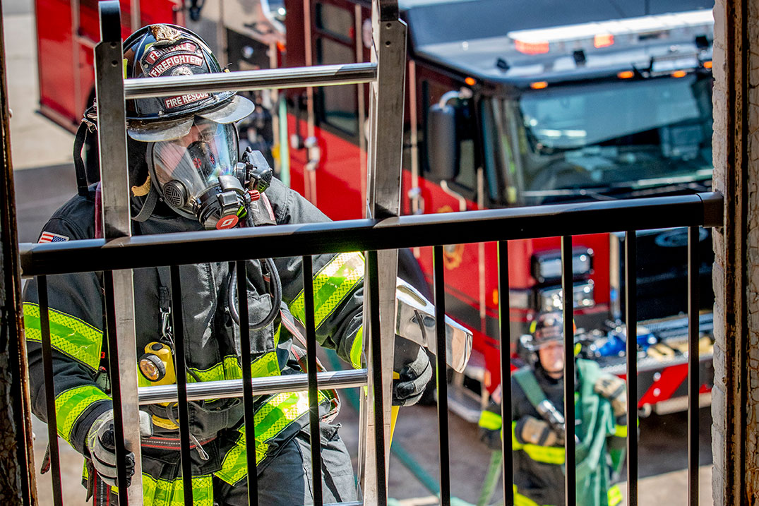 A firefighter in turnout gear climbing a ladder into a building and a firefighter carrying hose from a fire truck behind.