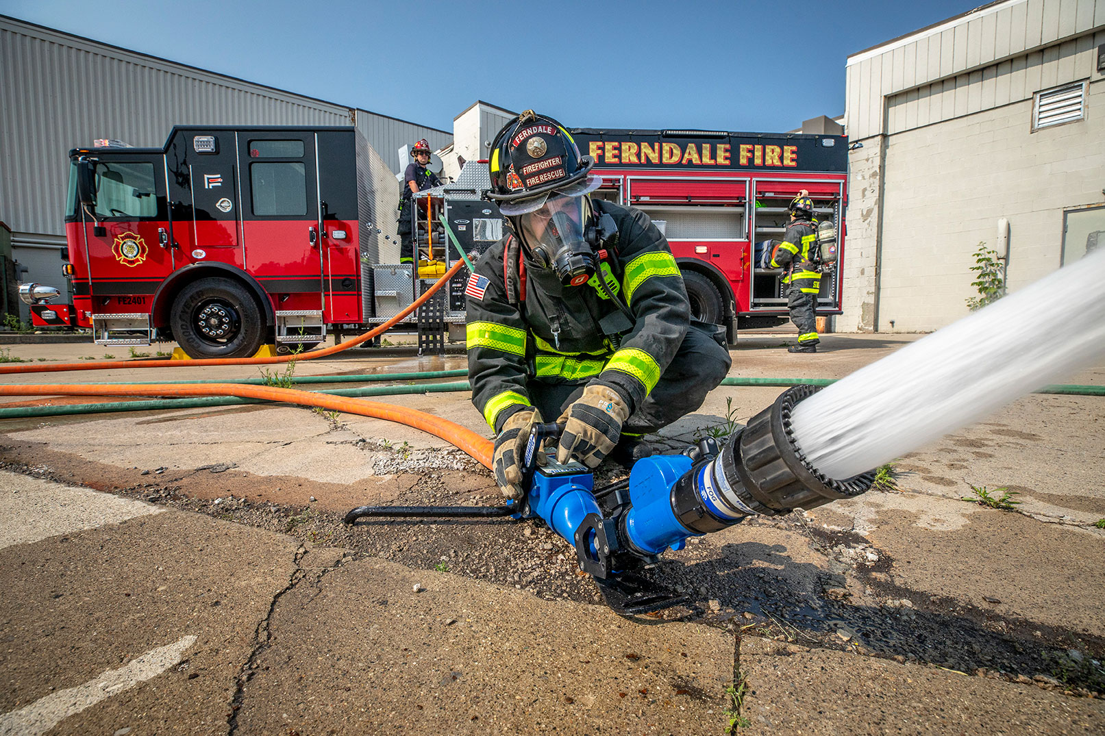 A firefighter in turnout gear spraying water from a hose with two other firefighters by a fire truck behind.