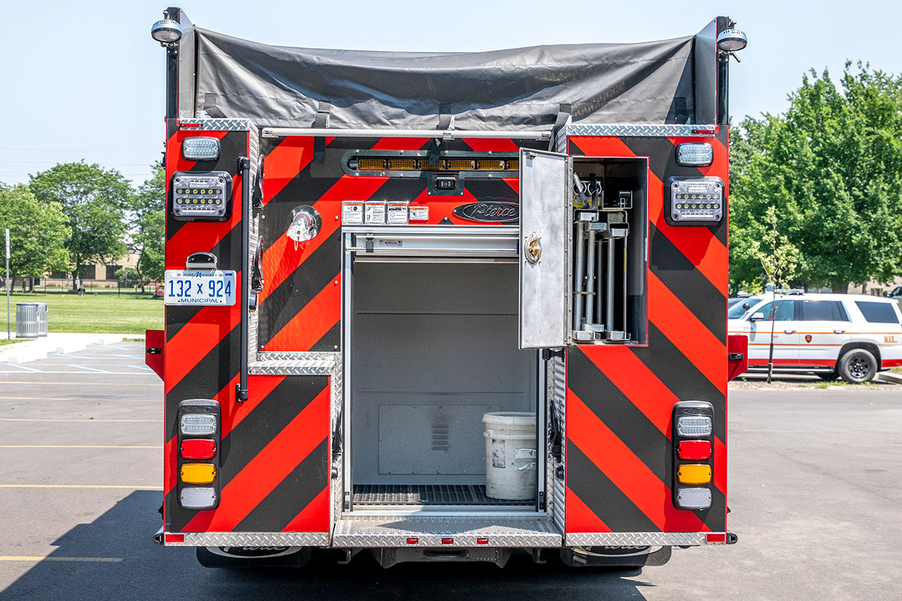 The rear of a fire truck with the compartments open showing ground ladders and a five gallon bucket.