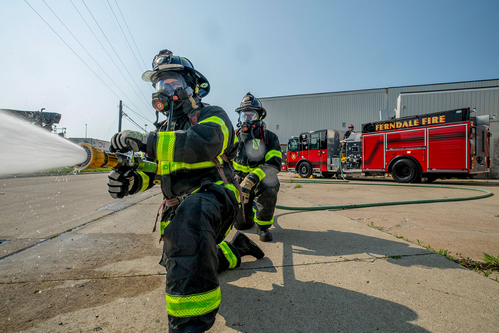 Two firefighters in turnout gear kneeling and spraying water from a hose and a firefighter operating a pump panel.