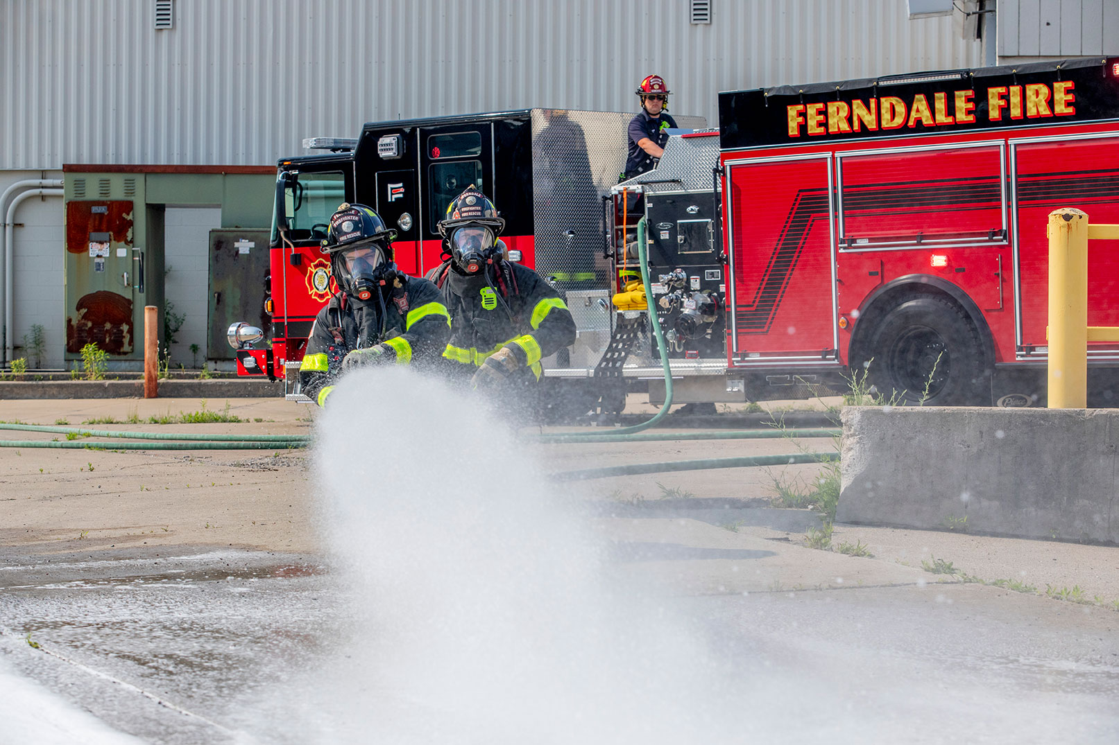 Two firefighters in turnout gear spraying water from a fire hose and a firefighter operating the pump.