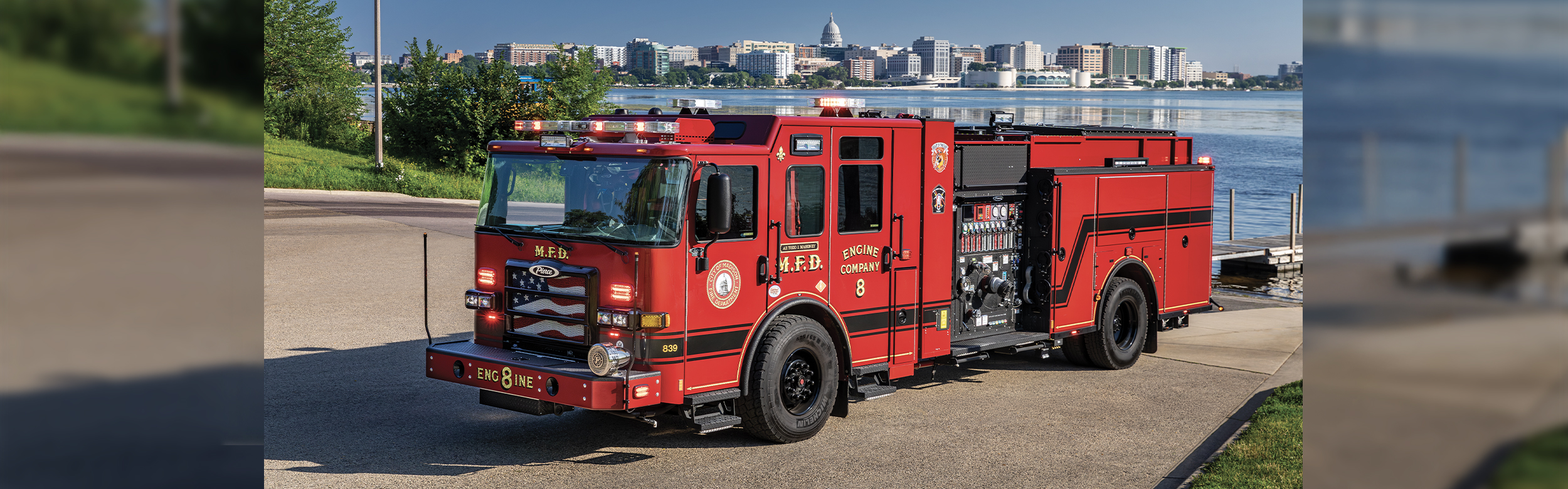 The driver's side of an Enforcer Volterra Electric Fire Truck in front of a lake and city skyline on a sunny day.