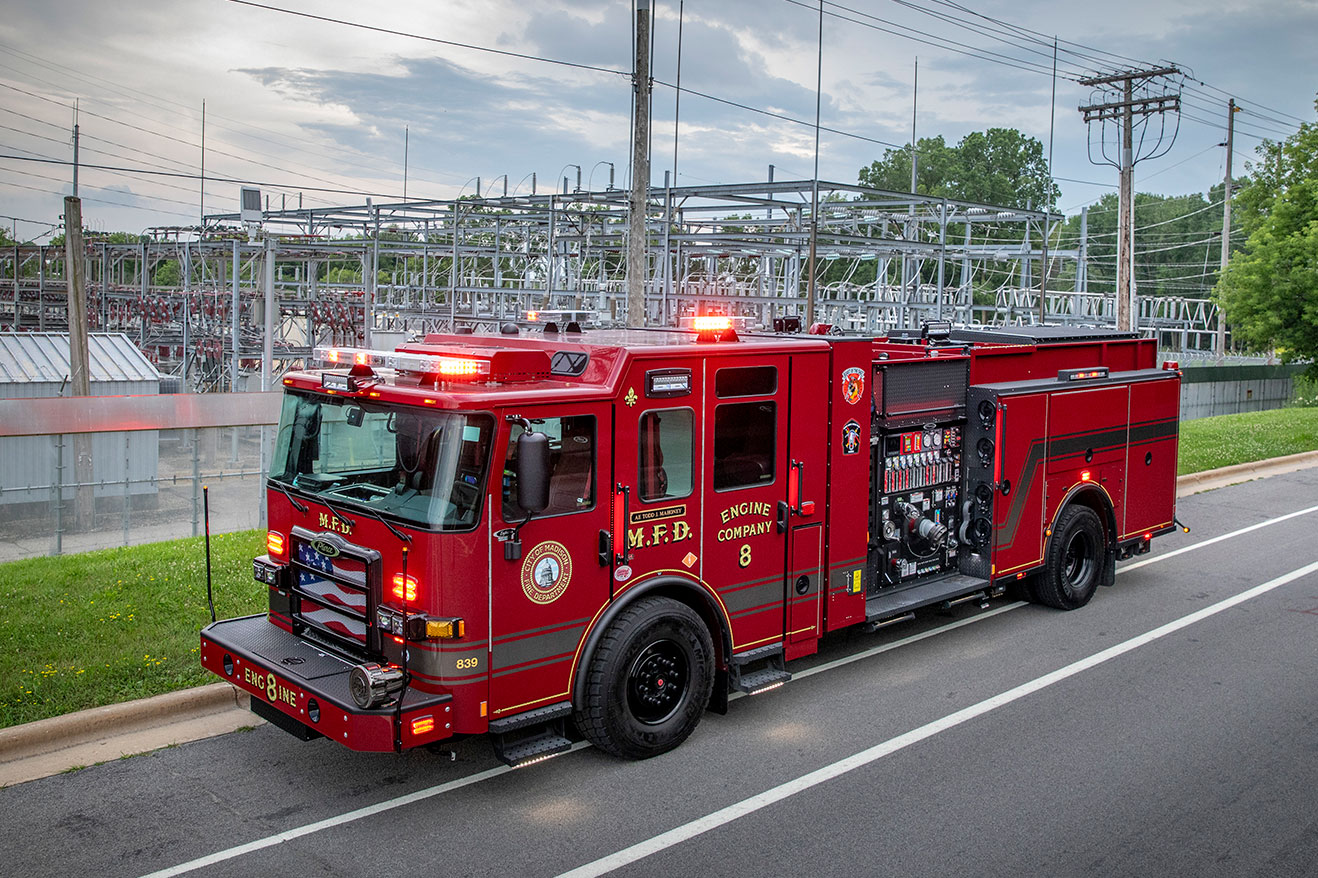 The drivers side of an Enforcer Volterra Electric Fire Truck in front of an industrial power facility on a cloudy day.