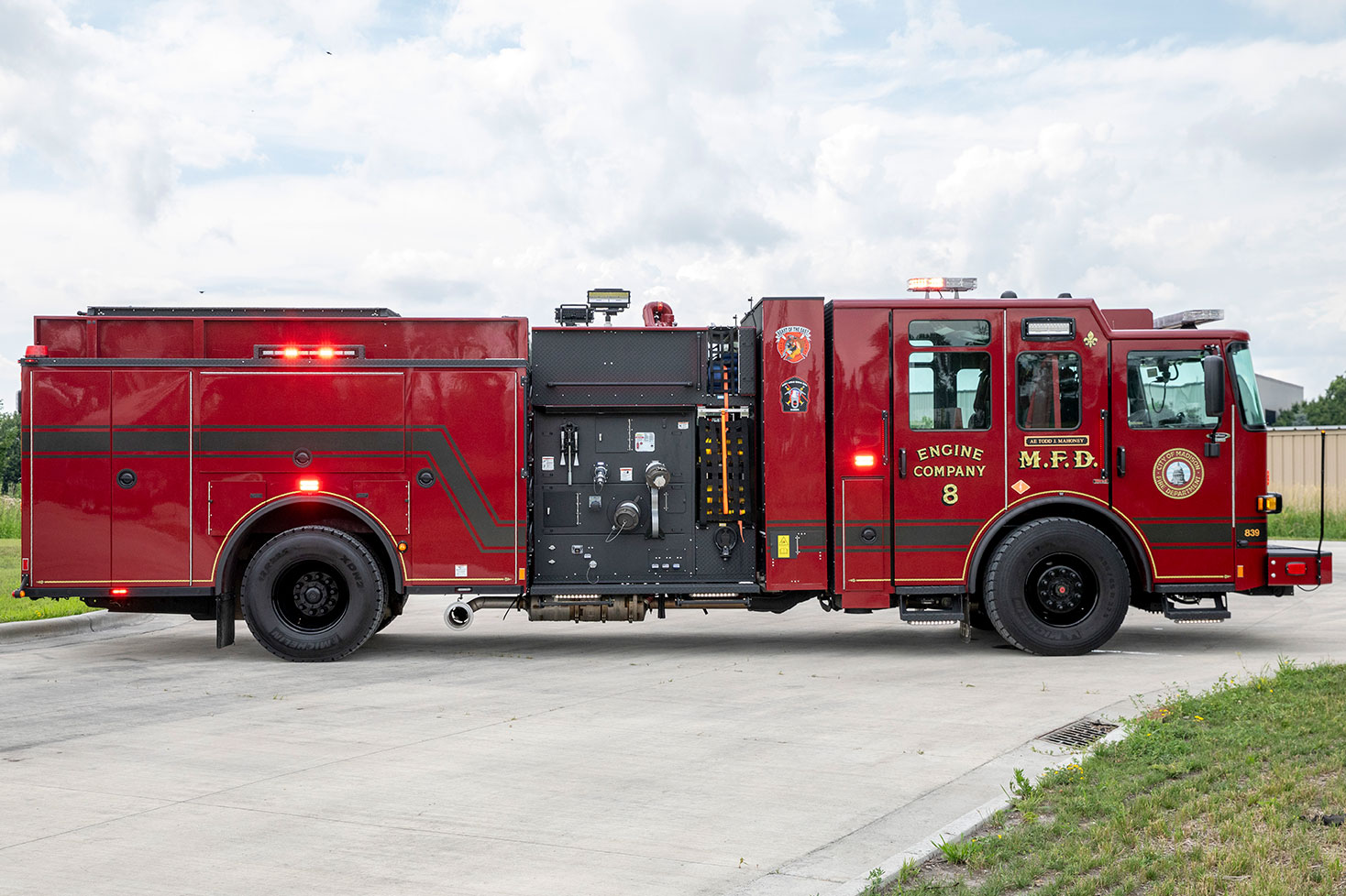 The officers side of an Enforcer Volterra Electric Fire Truck with the lights on in a parking lot on a cloudy day.
