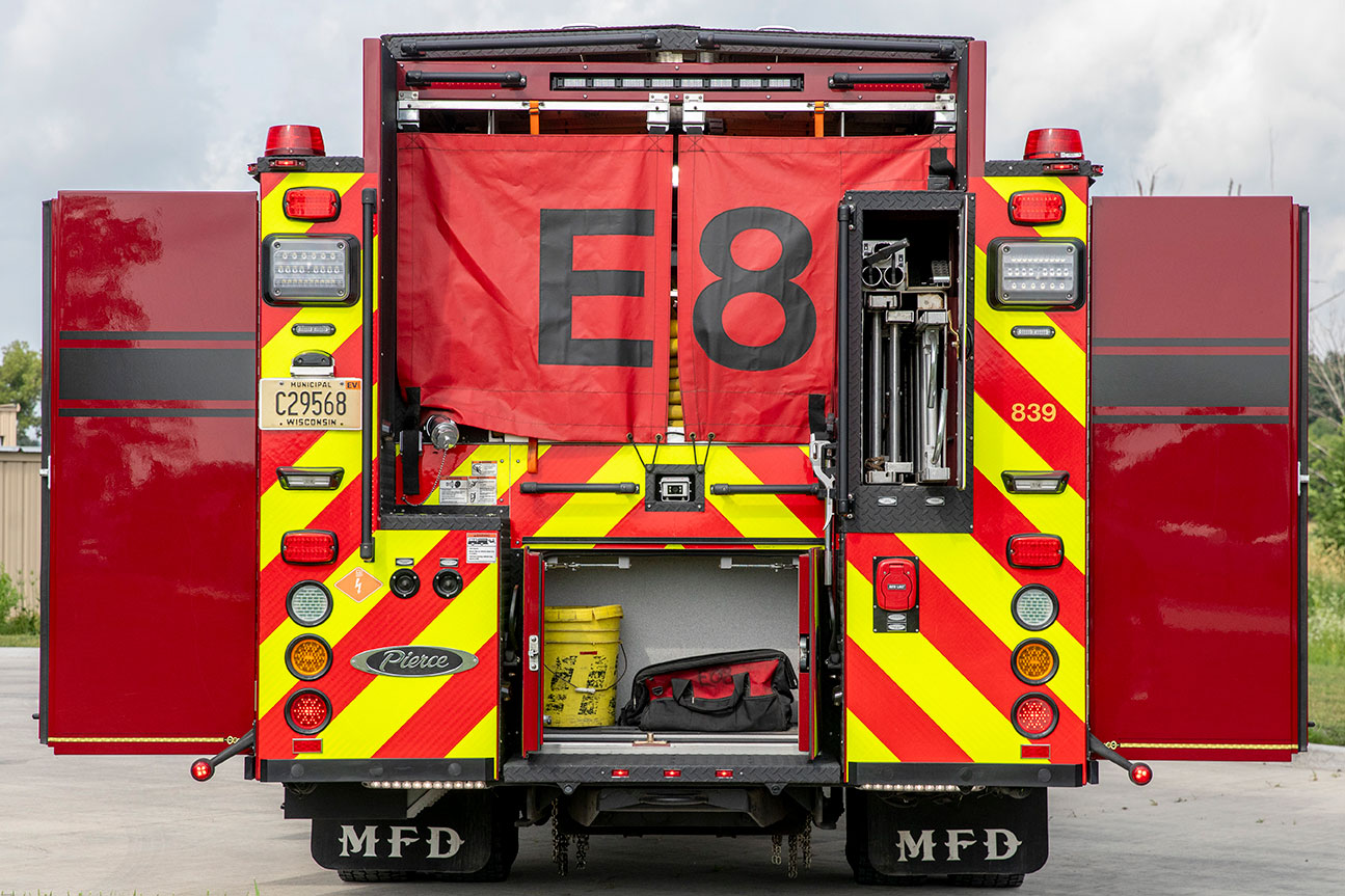 The rear of a pumper fire truck with a chevron pattern and the hose bed covered with a red hosebed cover and the compartments open.