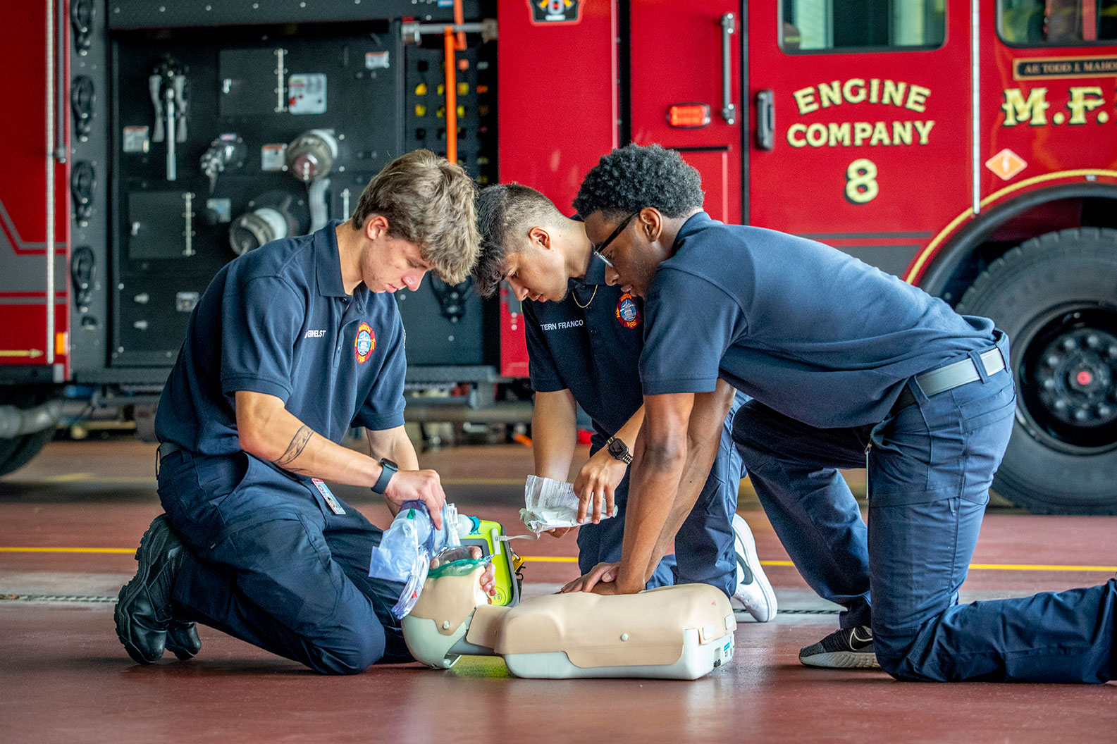 Three firefighters kneeling on the ground of a fire station practicing CPR on a mannequin with an electric fire truck in the background.