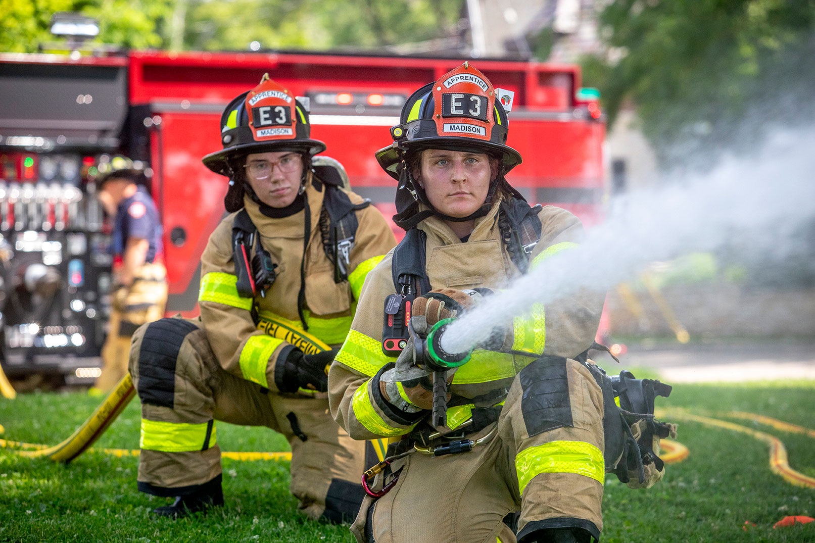 Two firefighters kneeling on the grass in turnout gear spraying water from a fire hose and another firefighter operating the pump.
