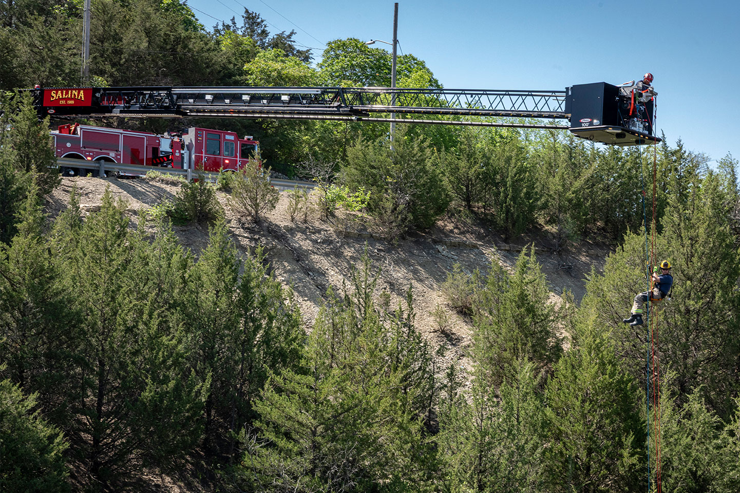 A firefighter hanging onto ropes from an aerial platform bucket over a cliff as the truck is parked on a road with the ladder extended.