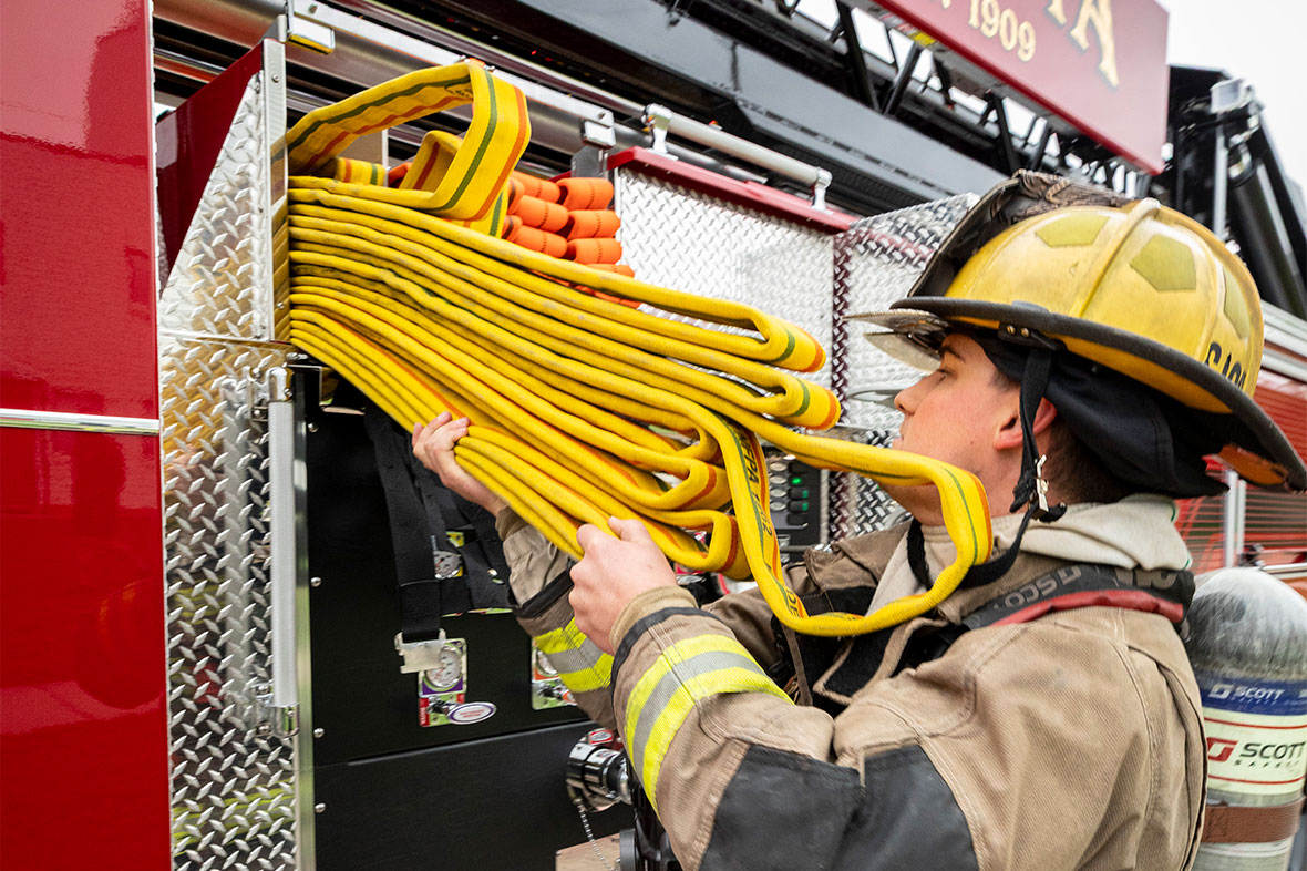 A firefighter in turnout gear pulling hose from the crosslays on the drivers side of a fire truck.