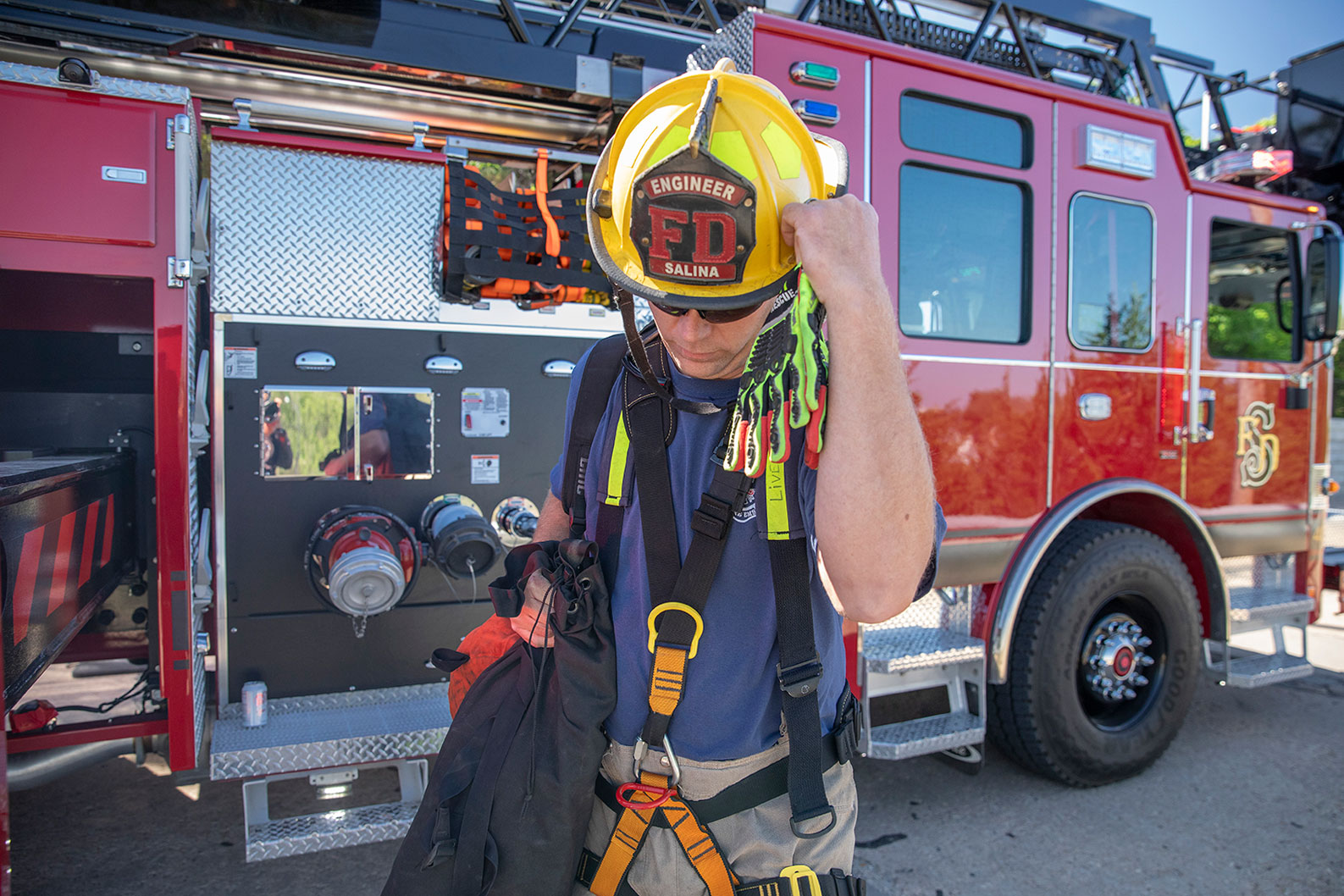 A firefighter with turnout gear on carrying gloves and a bag standing next to a fire truck.