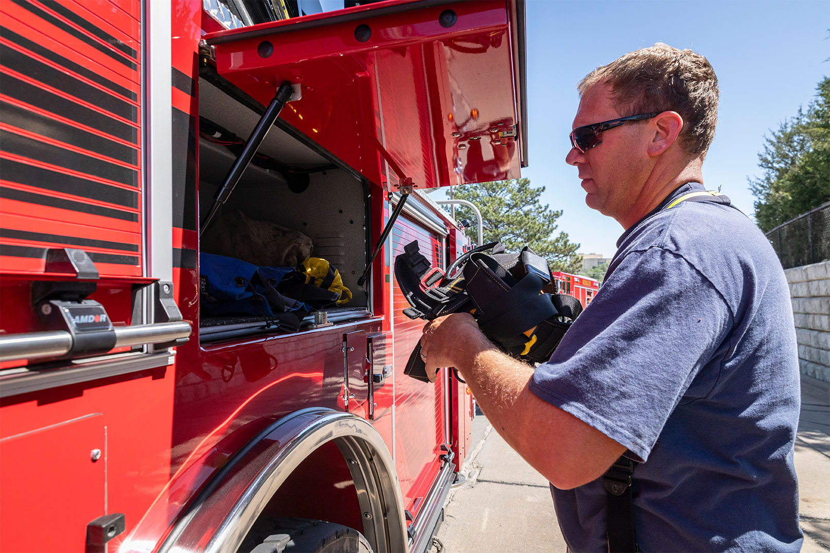A male firefighter grabbing equipment out of a compartment on the drivers side of a fire truck on a sunny day.