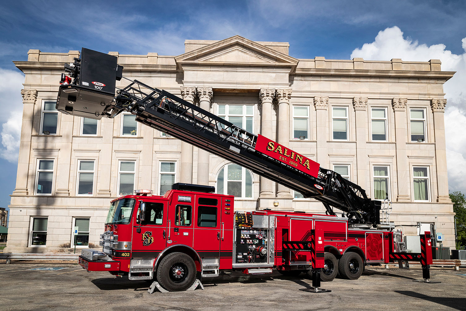 The drivers side of an Enforcer 100 Heavy-Duty Steel Aerial Platform fire truck in front of a historic building with the aerial up.