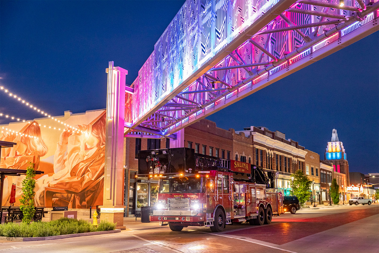 The front officers side of an aerial platform fire truck driving down a city street under a colorful lit bridge.