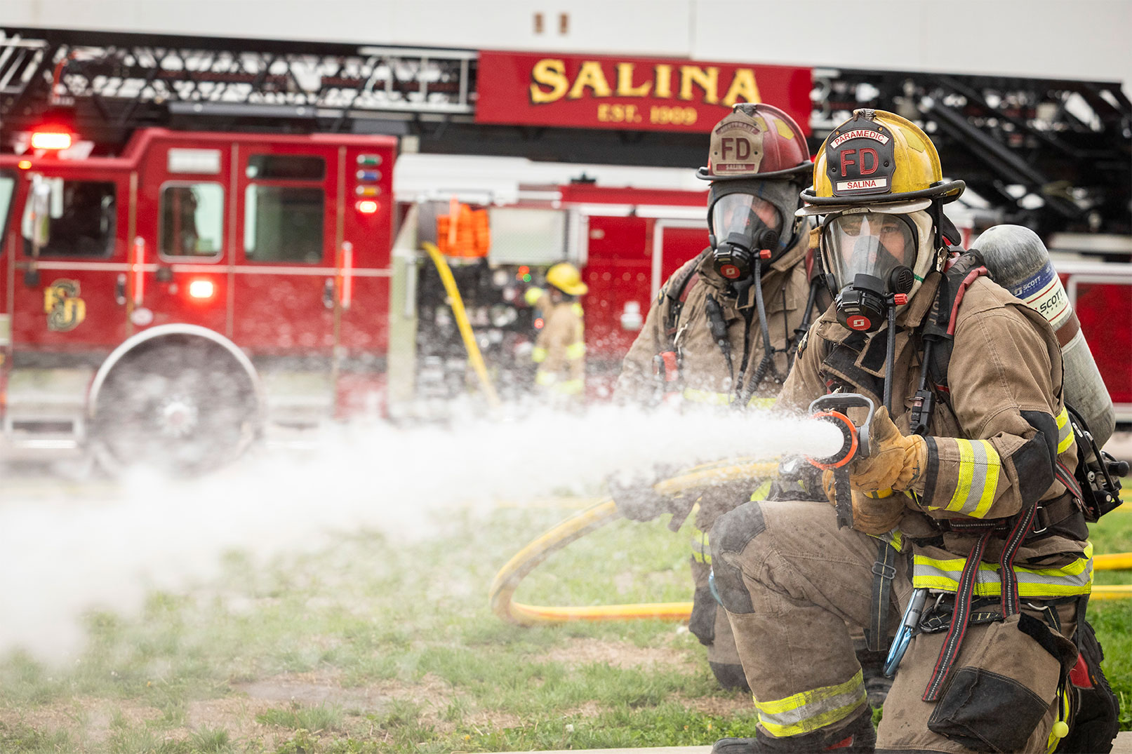 Two firefighters in turnout gear spraying water from a fire hose and a firefighter operating the pump panel.