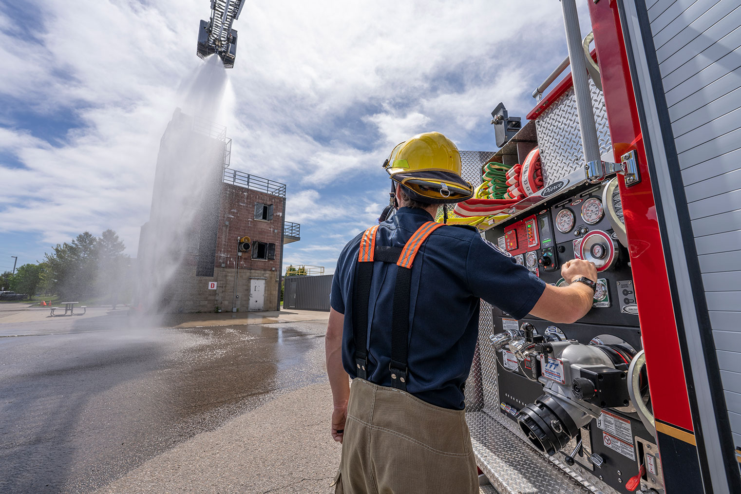 A firefighter with turnout gear on operating the pump of an aerial fire truck with water spraying from the aerial waterway.