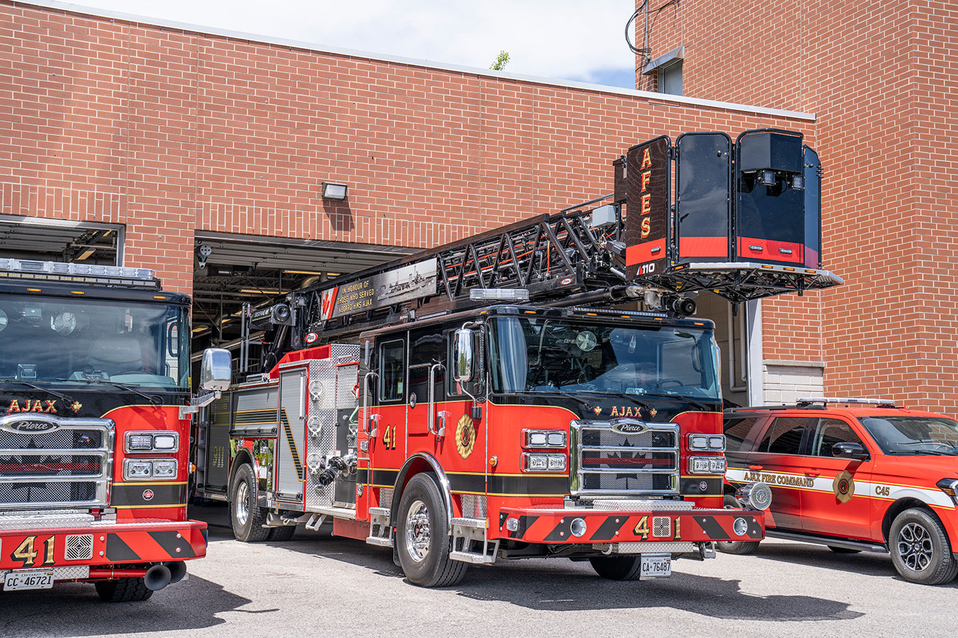 The front officers side of an Enforcer 110 Ascendant Aerial Platform parked in front of a fire station next to a pumper and car.