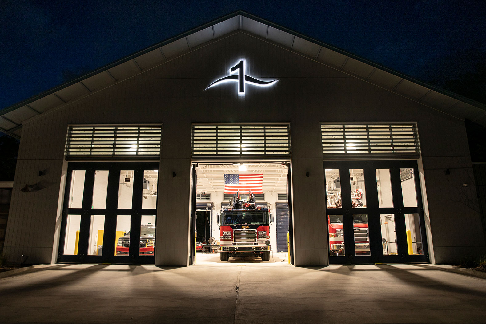 A fire station with three apparatus bays including two fire trucks and a chief car with one glass bay door open at night.
