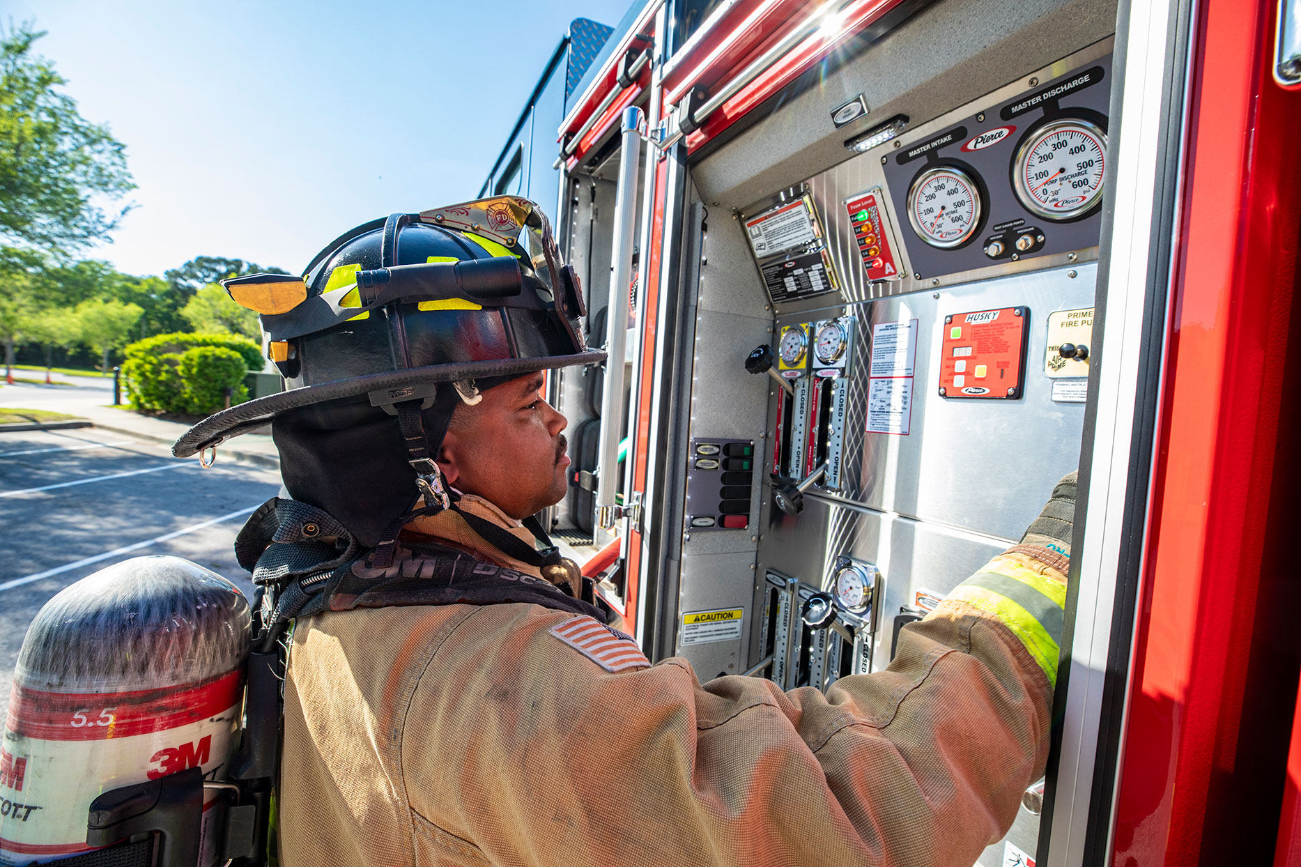 A firefighter in turnout gear operating a PUC pump on an aerial fire truck with buttons and levers in a parking lot on a sunny day.