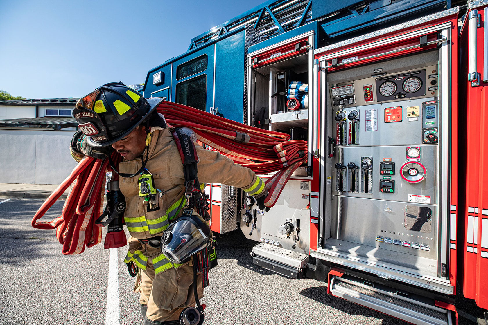 A firefighter in turnout gear pulling red fire hose from the crosslays of an Aerial fire truck in a parking lot.