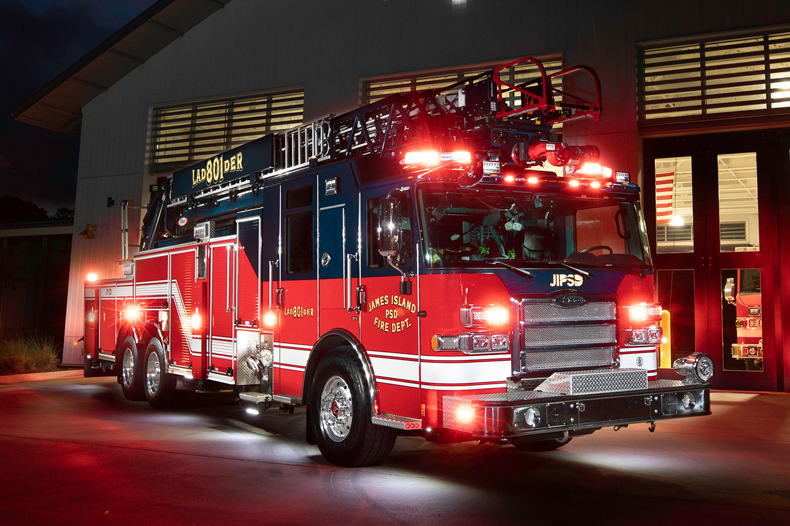 The officers side of an Aerial fire truck with the lights on parked in front of a fire station at night.