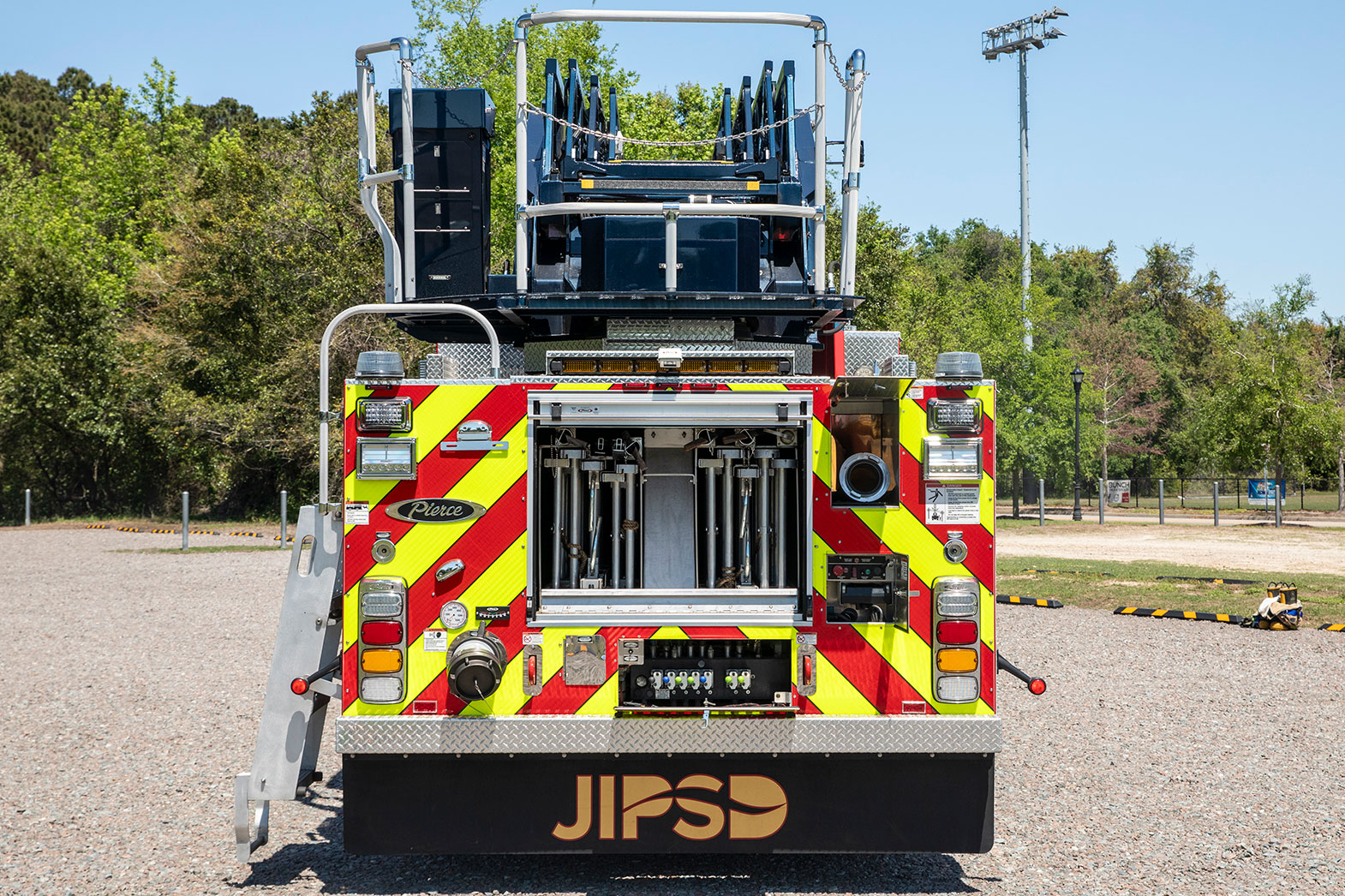 The rear of an Aerial fire truck parked in a gravel parking lot with the rear compartment open showing ground ladders inside.
