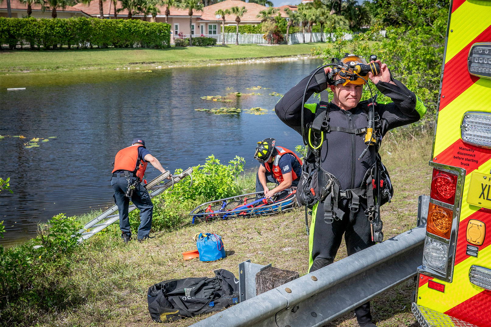 A firefighter in a dive suit putting on a breathing mask beside a fire truck and two firefighters in life vests behind by the lake.