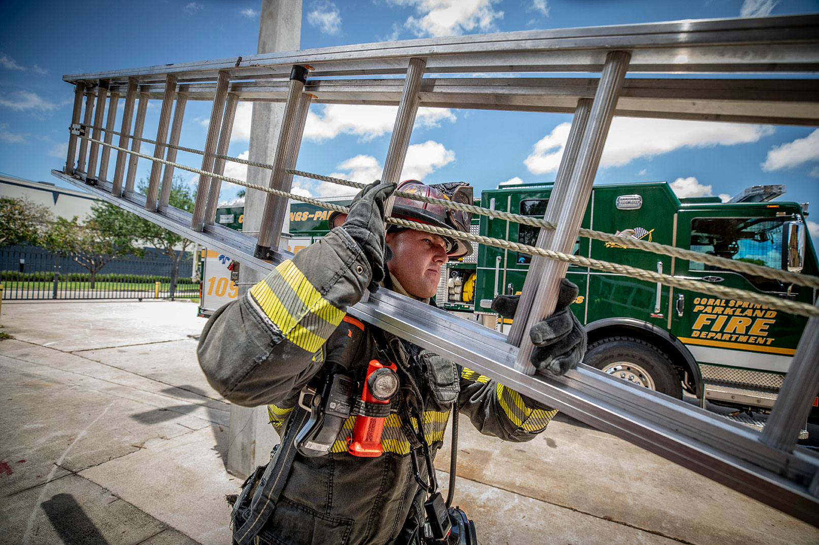 A firefighter in turnout gear carrying a ground ladder on his shoulder with a green fire truck in the background on a sunny day.