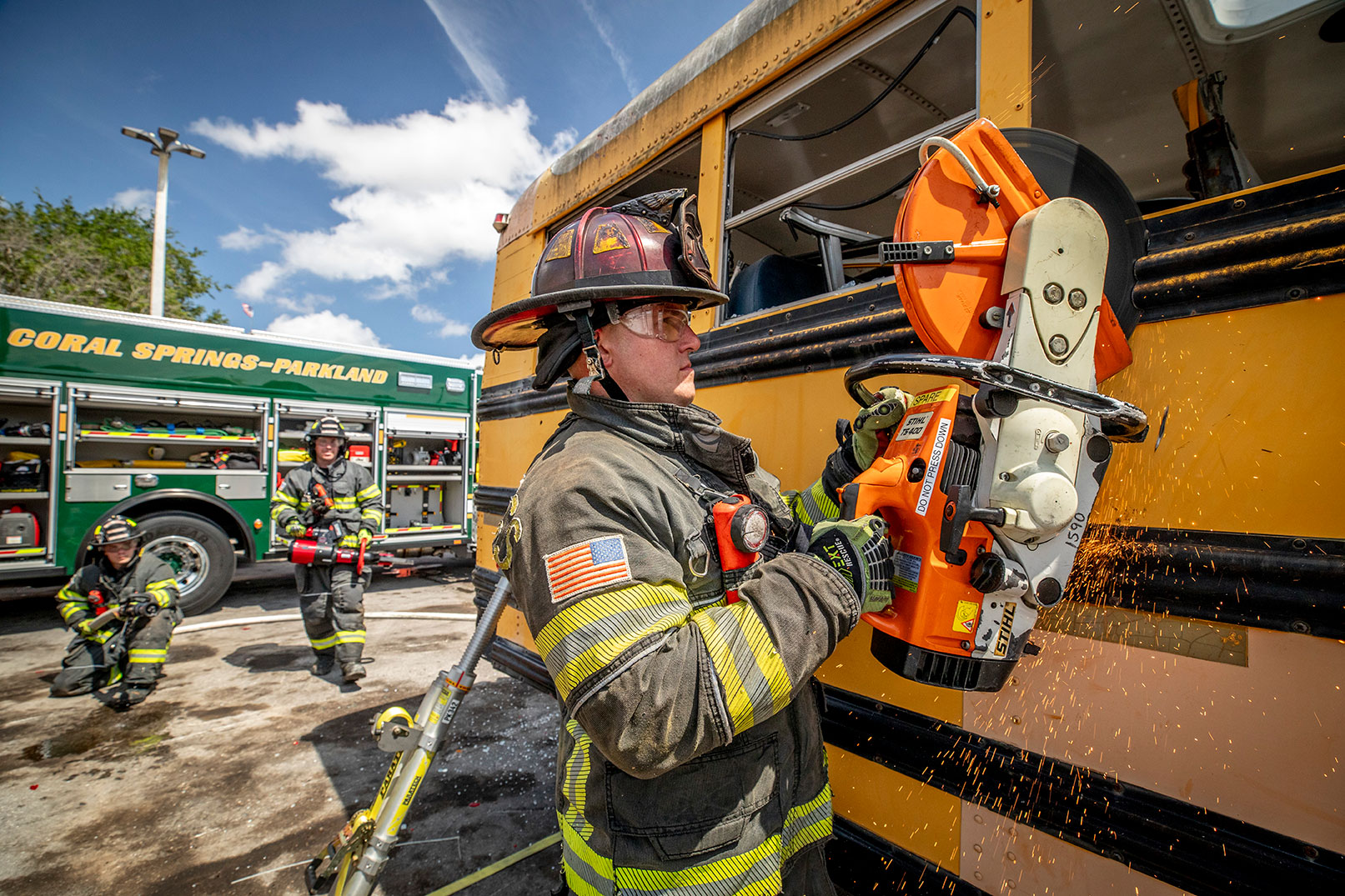 A firefighter in turnout gear using a saw to cut into the side of a school bus with two firefighters by a fire truck behind.