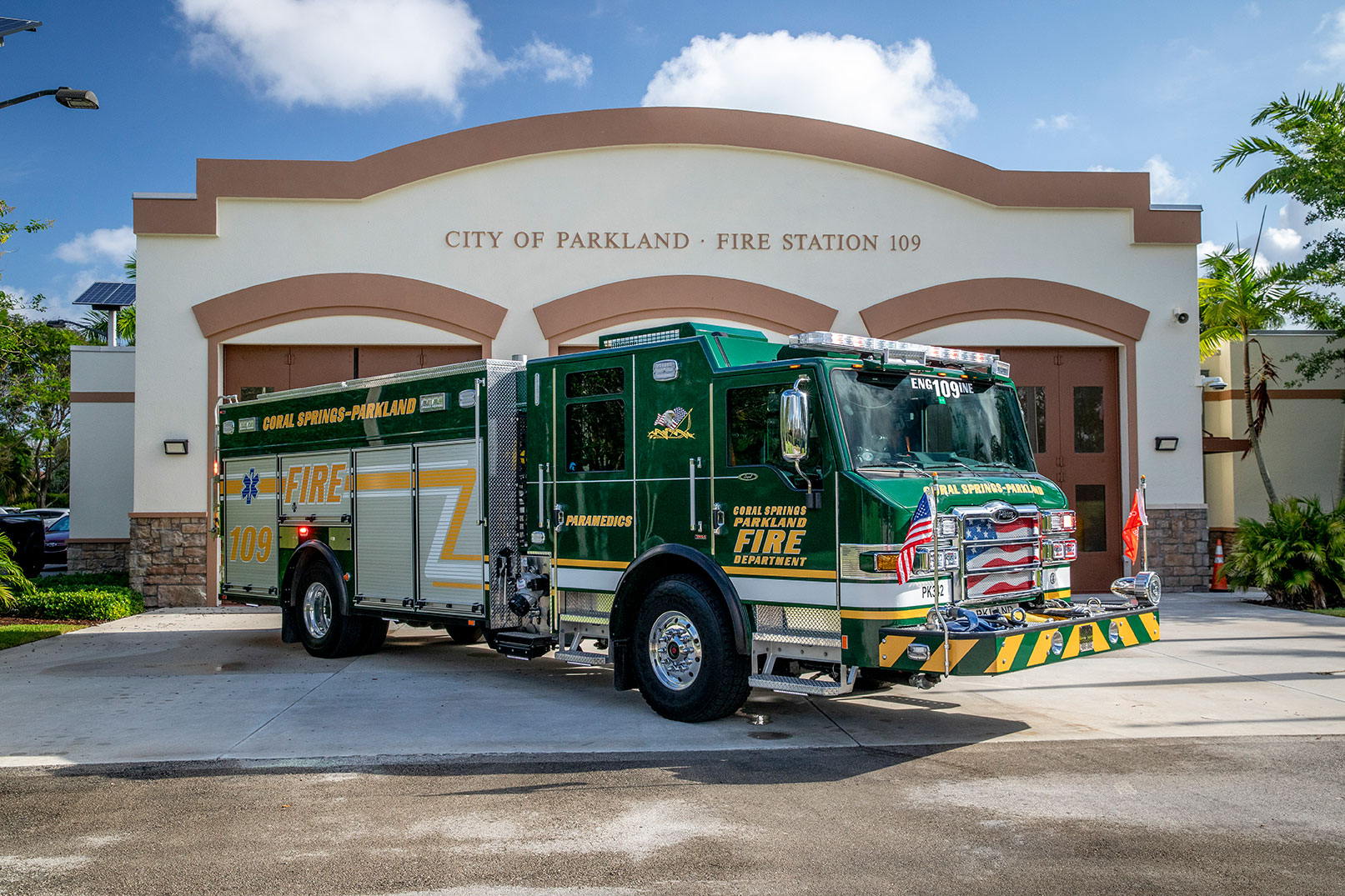 The front officers side of an Impel PUC Pumper parked in front of a fire station on a sunny day with palm trees.