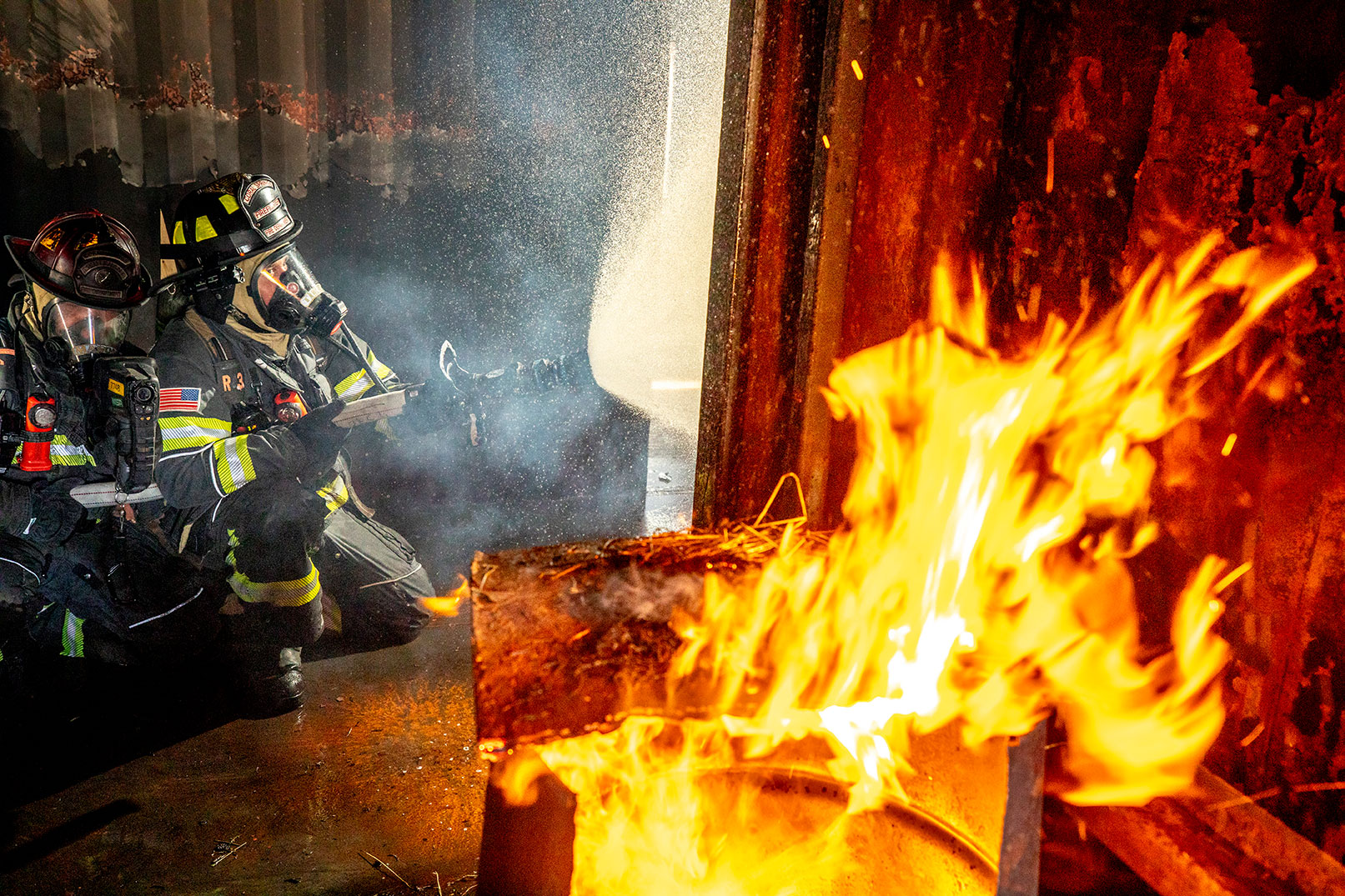 Two firefighters kneeling on the ground spraying water from a fire hose in a steel building with a fire in the corner.