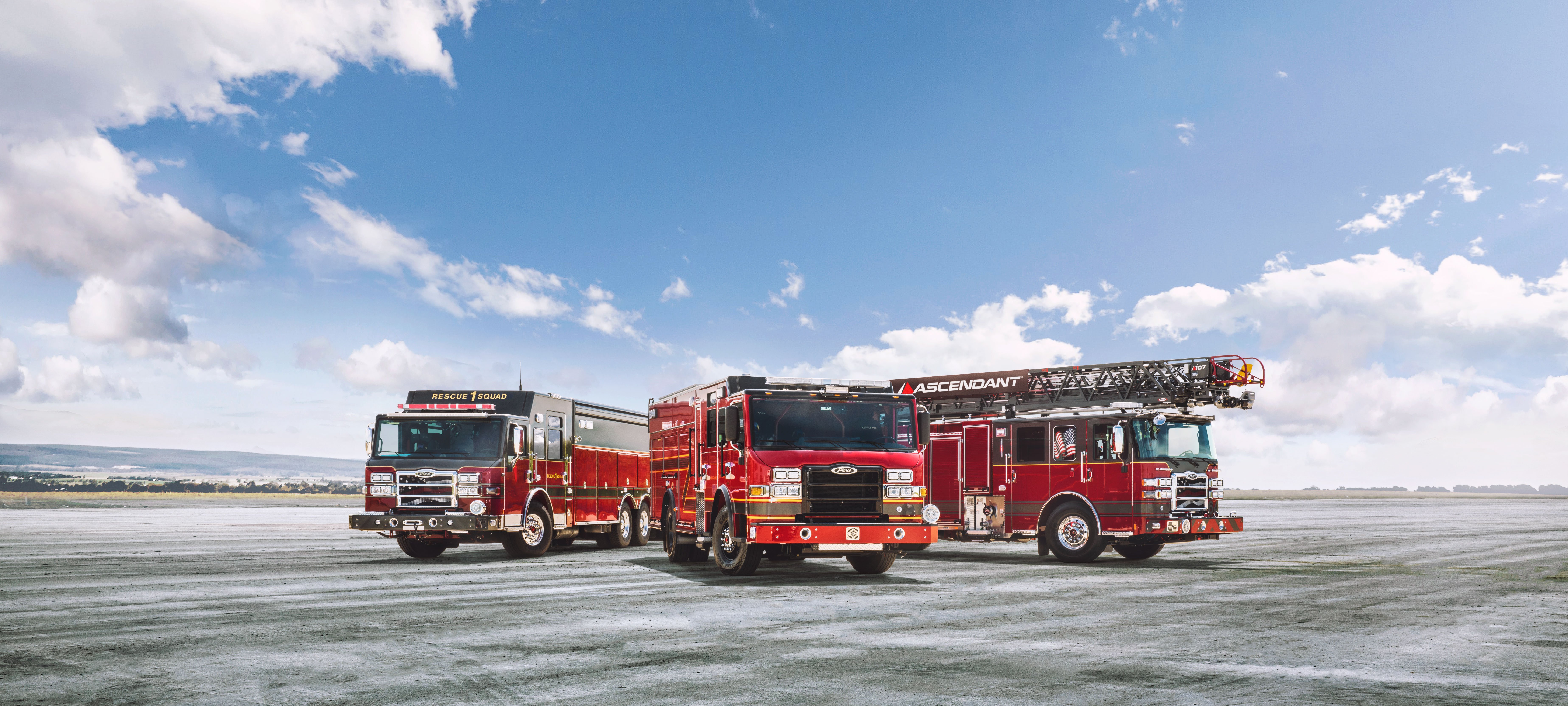 Three custom Pierce fire trucks parked outside on open terrain near water. 