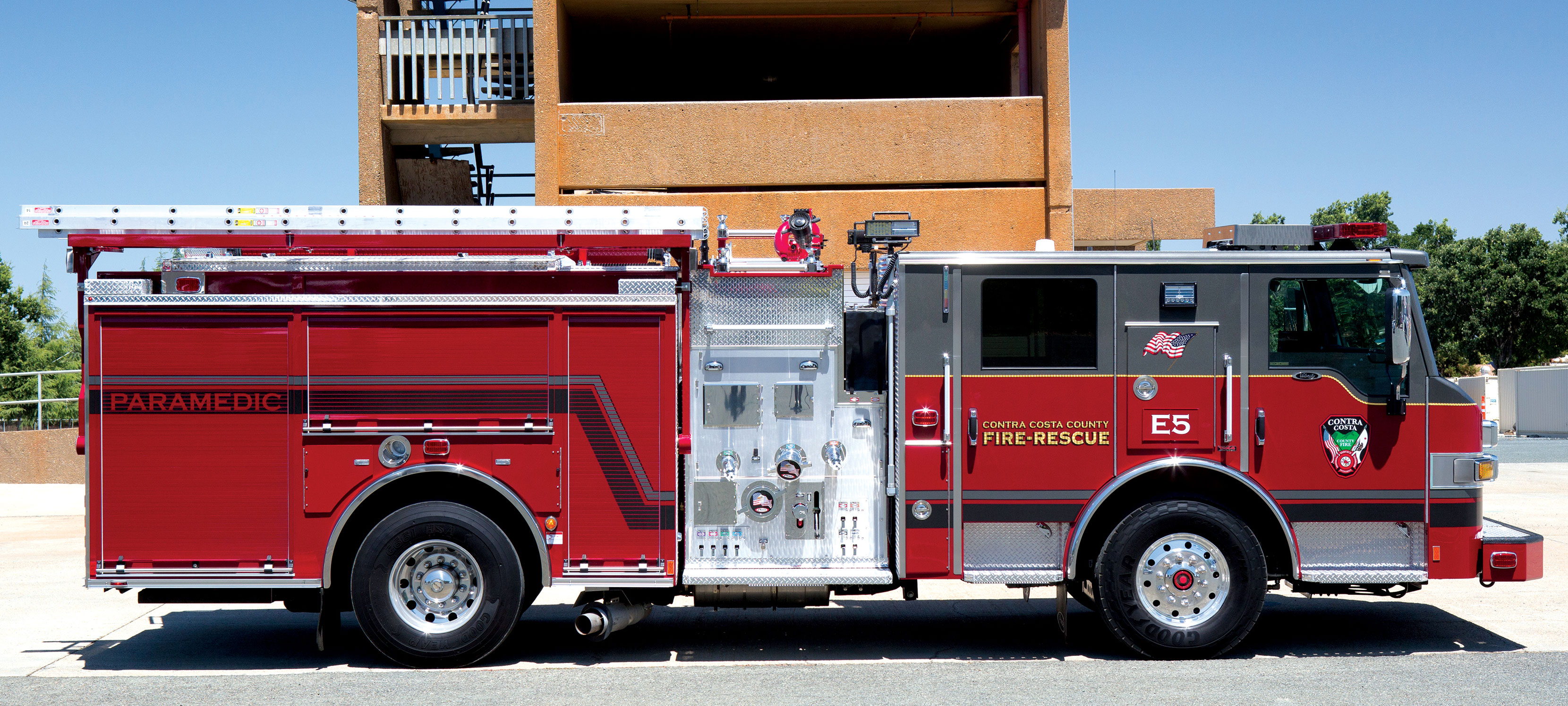 Low Crosslays on the officer’s side of a Pierce Fire Truck parked outside near a building on a sunny day. 