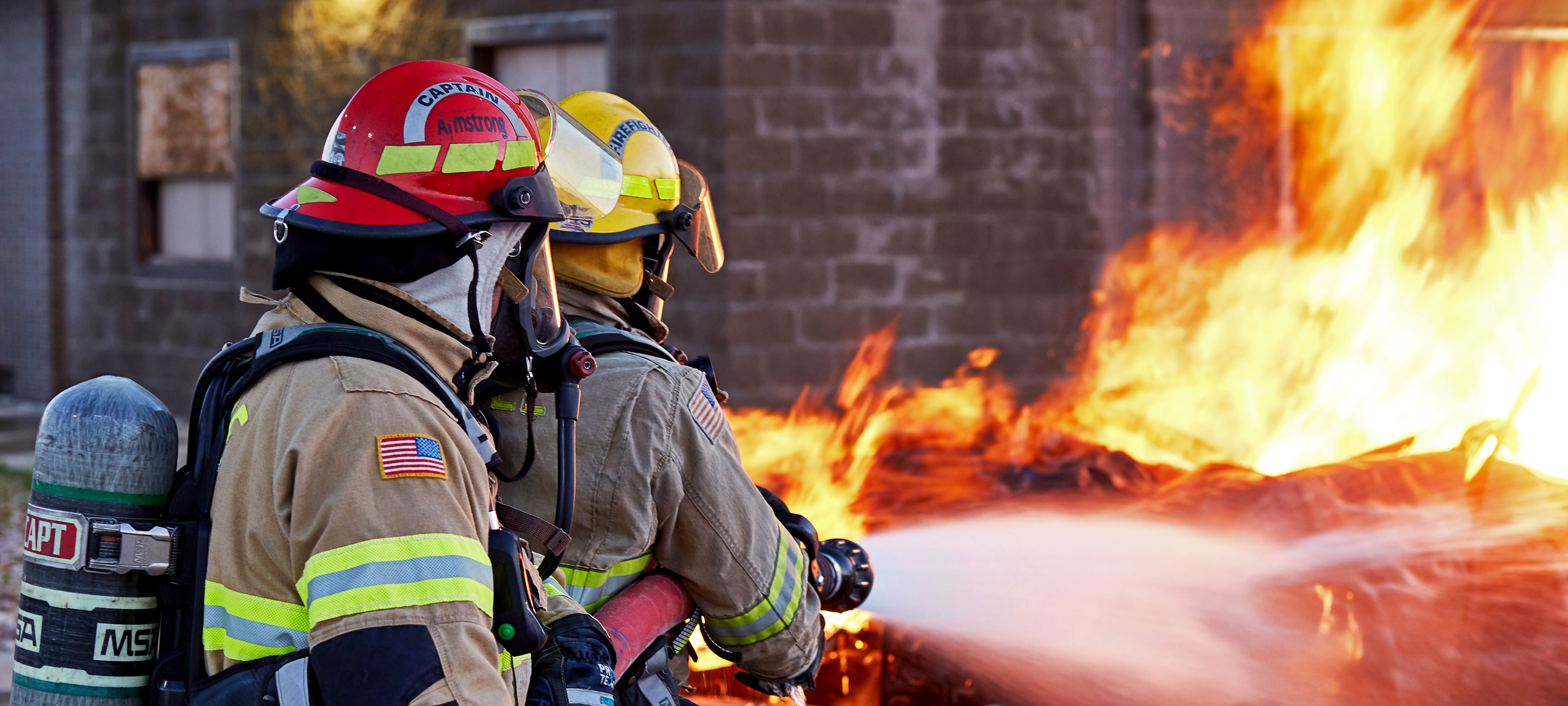 Two firefighters pumping water through a hose on a fire near a building. 