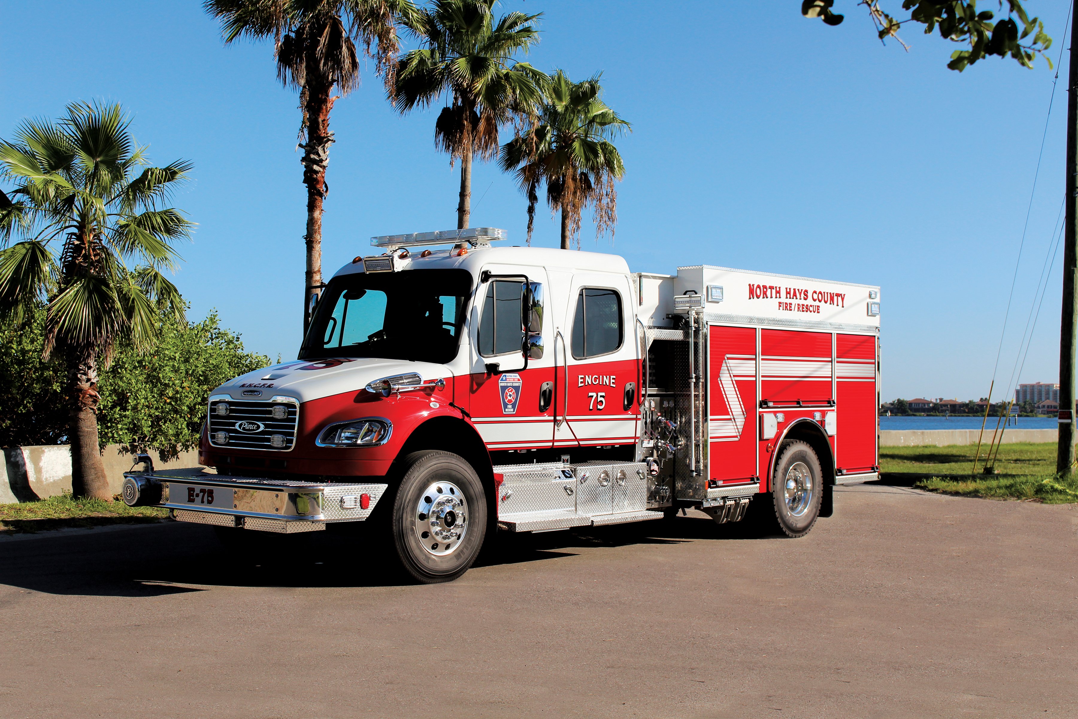 Drivers side of a Pierce Freightliner commercial chassis fire truck parked outside surrounded by trees. 