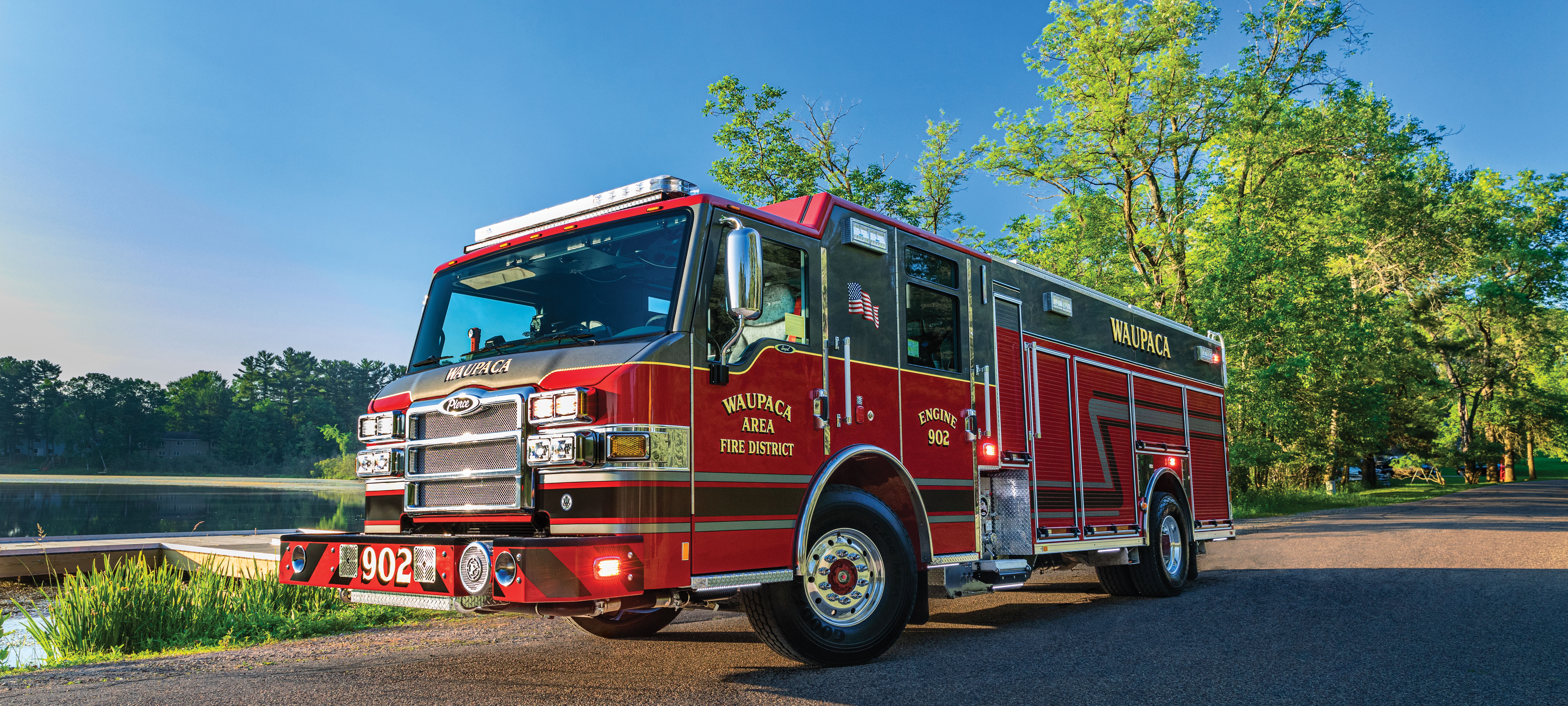 Pierce Impel custom fire truck chassis parked outside near trees beside the water on a sunny day. 