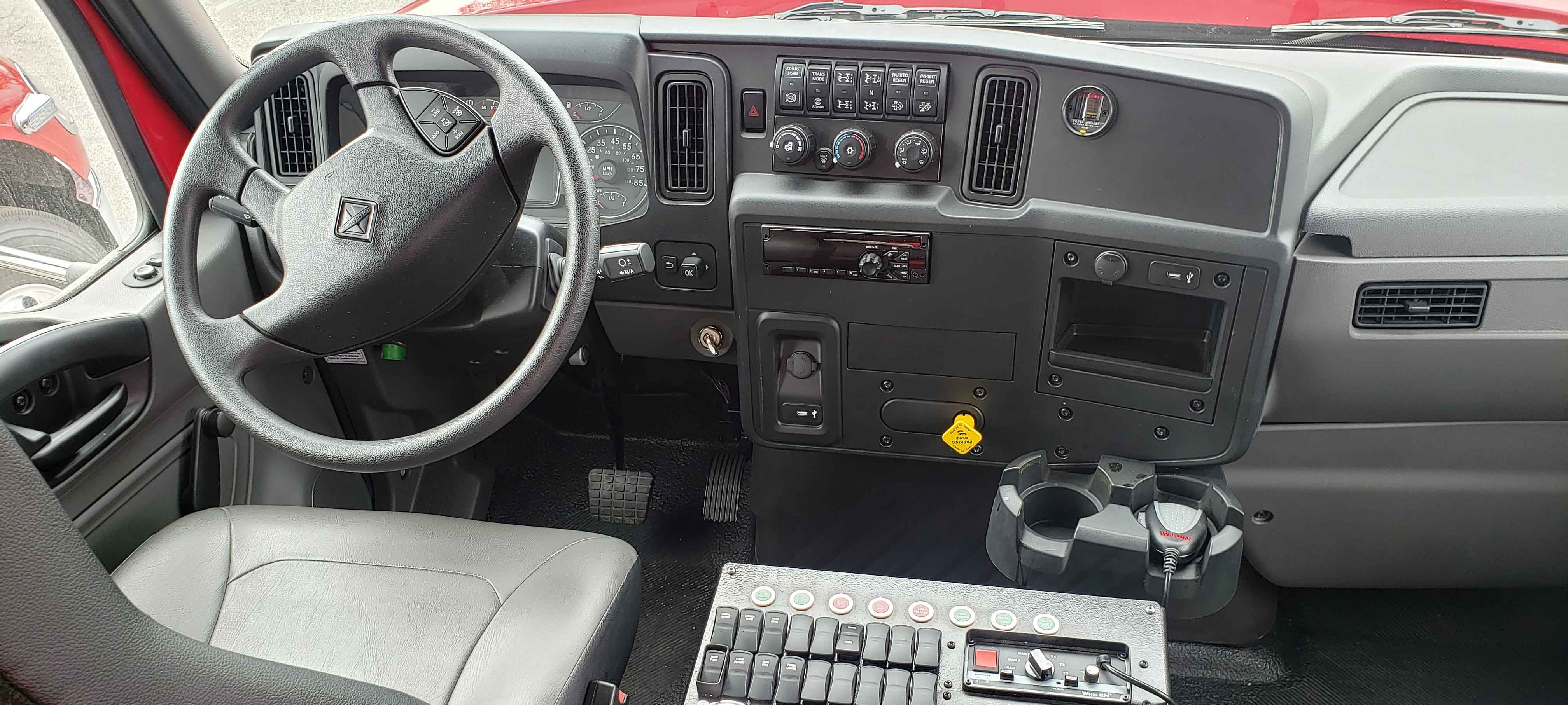 Grey steering wheel, drivers side seat and radio controls on the interior of a Pierce International Commercial fire truck chassis cab interior. 