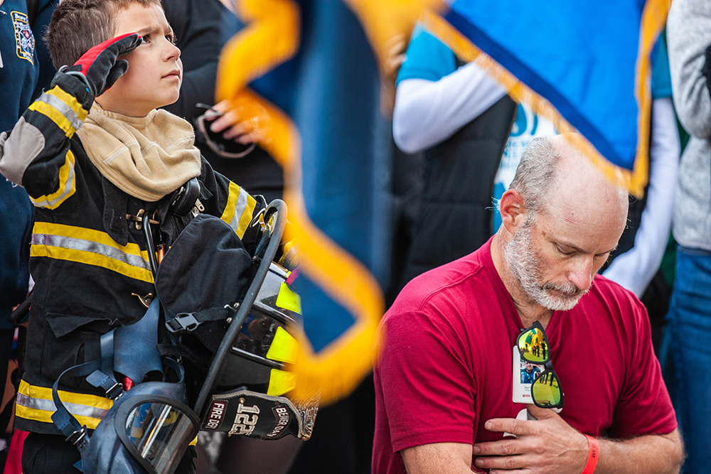 A boy in turnout gear standing for the National Anthem next to a man sitting with his hand on his chest at Lambeau Field for the Pierce Manufacturing 9/11 Stair Climb Event.