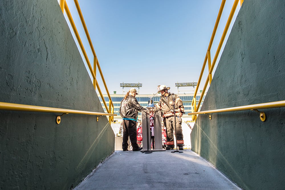 A Firefighter in turnout gear and a man ringing the bell at the entrance of the tunnel at Lambeau Field at the Pierce Manufacturing 9/11 Stair Climb Event.