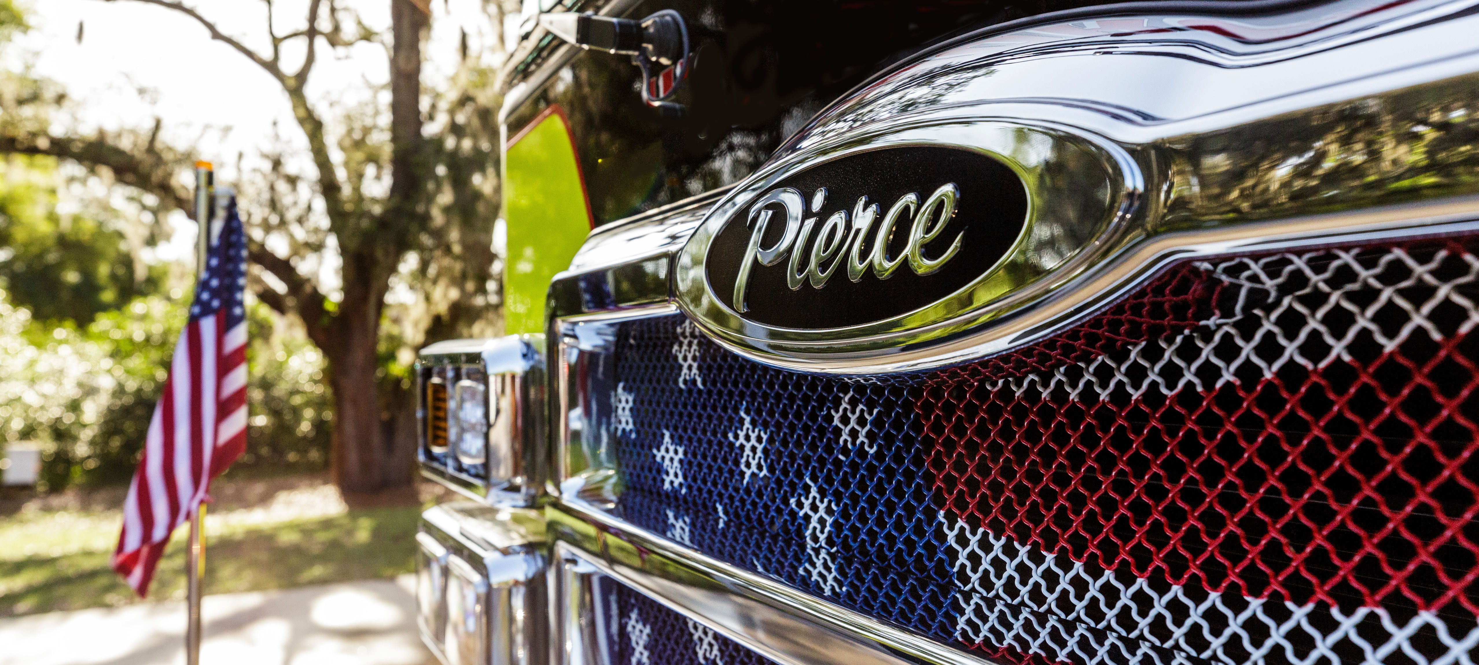Pierce Fire Truck parked outside on a sunny day near an American flag with a close up of the front bumper logo.