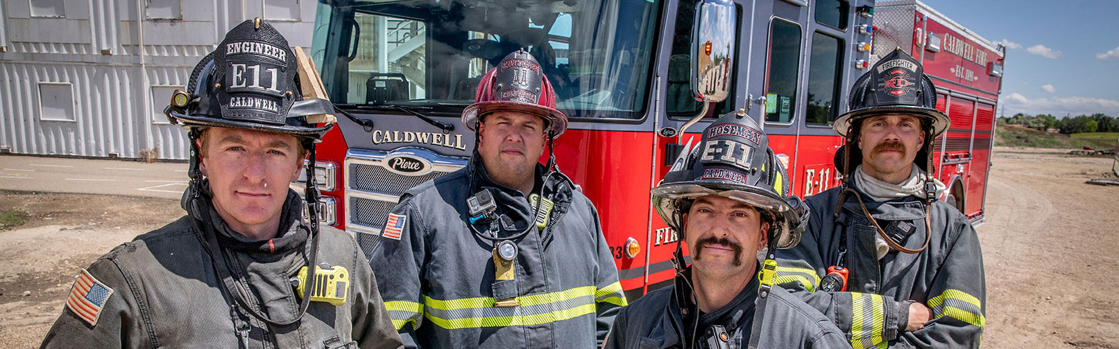 Four-firefighters-in-turnout-gear-standing-in-front-of-a-fire-truck-at-a-firefighter-training-center