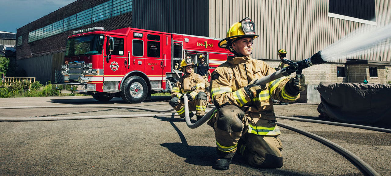 Firefighters pumping water from a Pierce PUC™ Fire Truck parked outside near a building on a sunny day. 