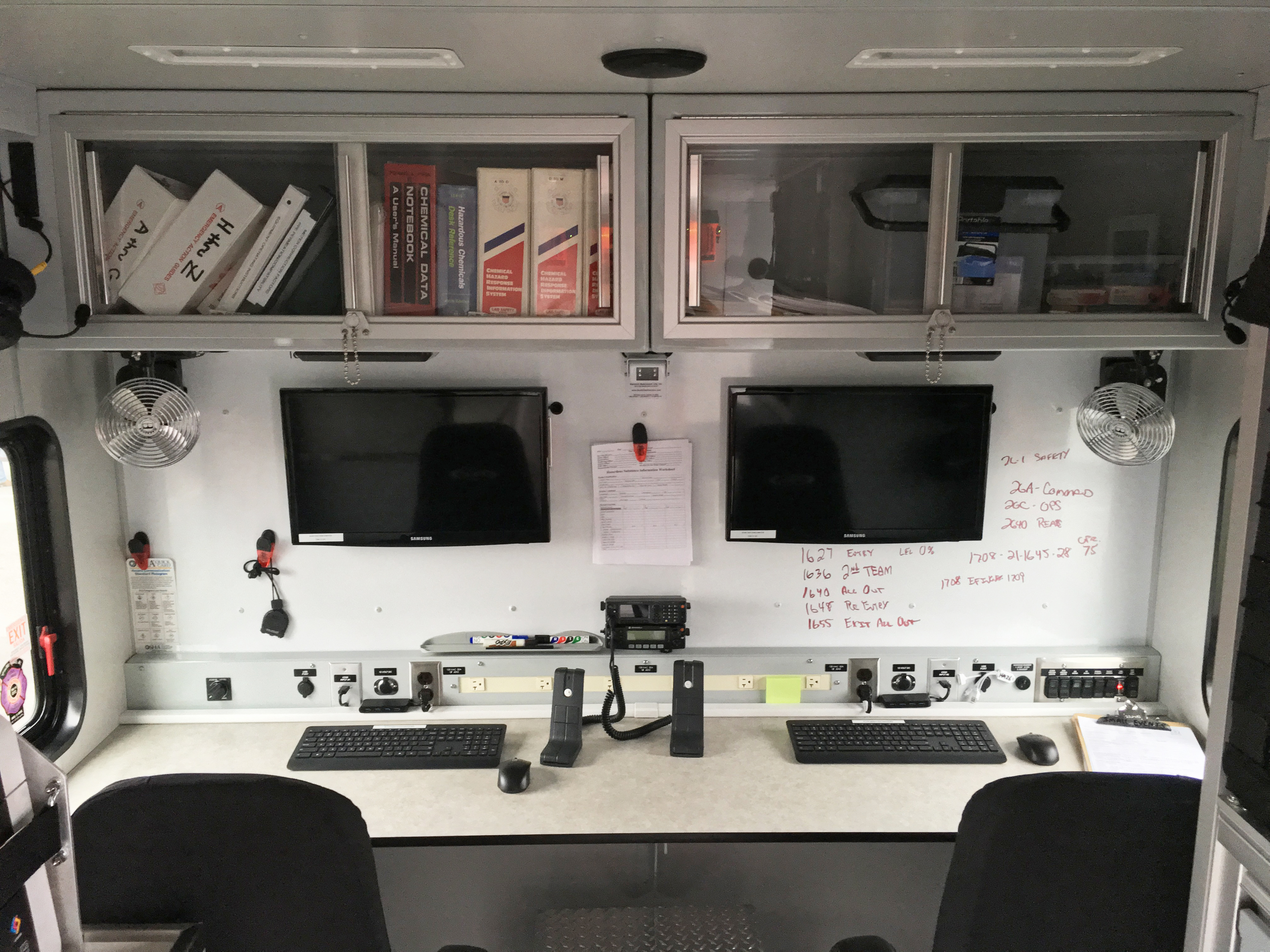Two workstations on the interior of a Pierce Combination Rescue Fire Truck with two computers, two chairs and shelving up above. 