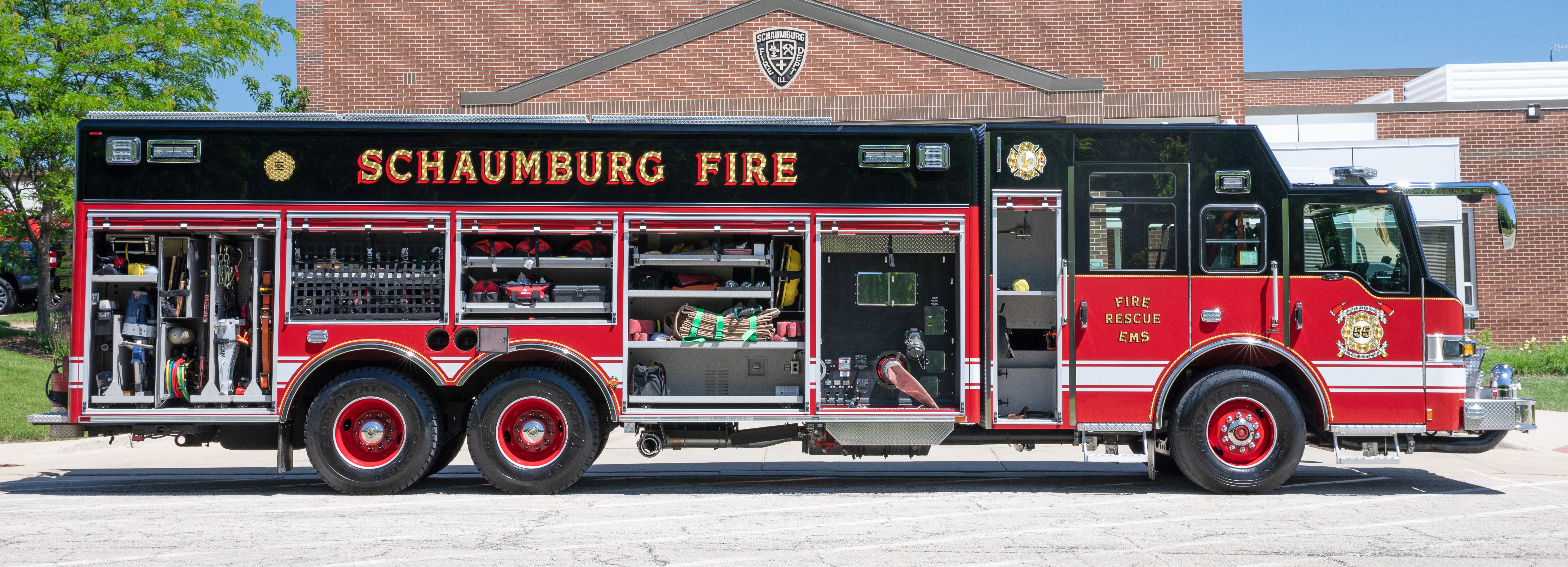 Driver’s side of a Pierce Non-Walk-In Rescue Fire Truck parked outside in front of a fire station on a sunny day with side compartments open showing storage space.  